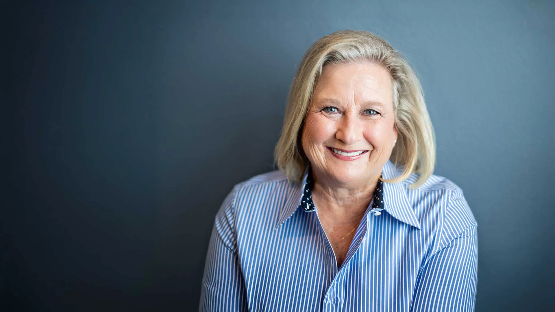 Smiling mature woman with blonde hair wearing blue striped shirt against a dark gray background.