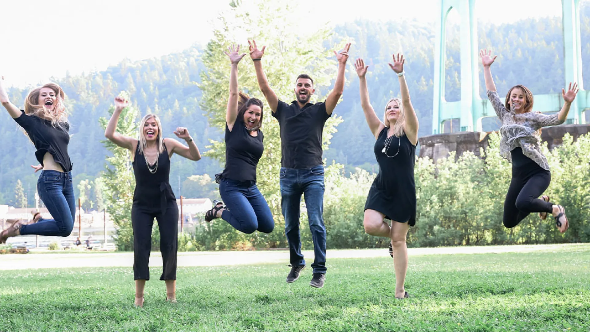 A group of six people joyfully jumping in the air outdoors, with greenery and a bridge in the background.