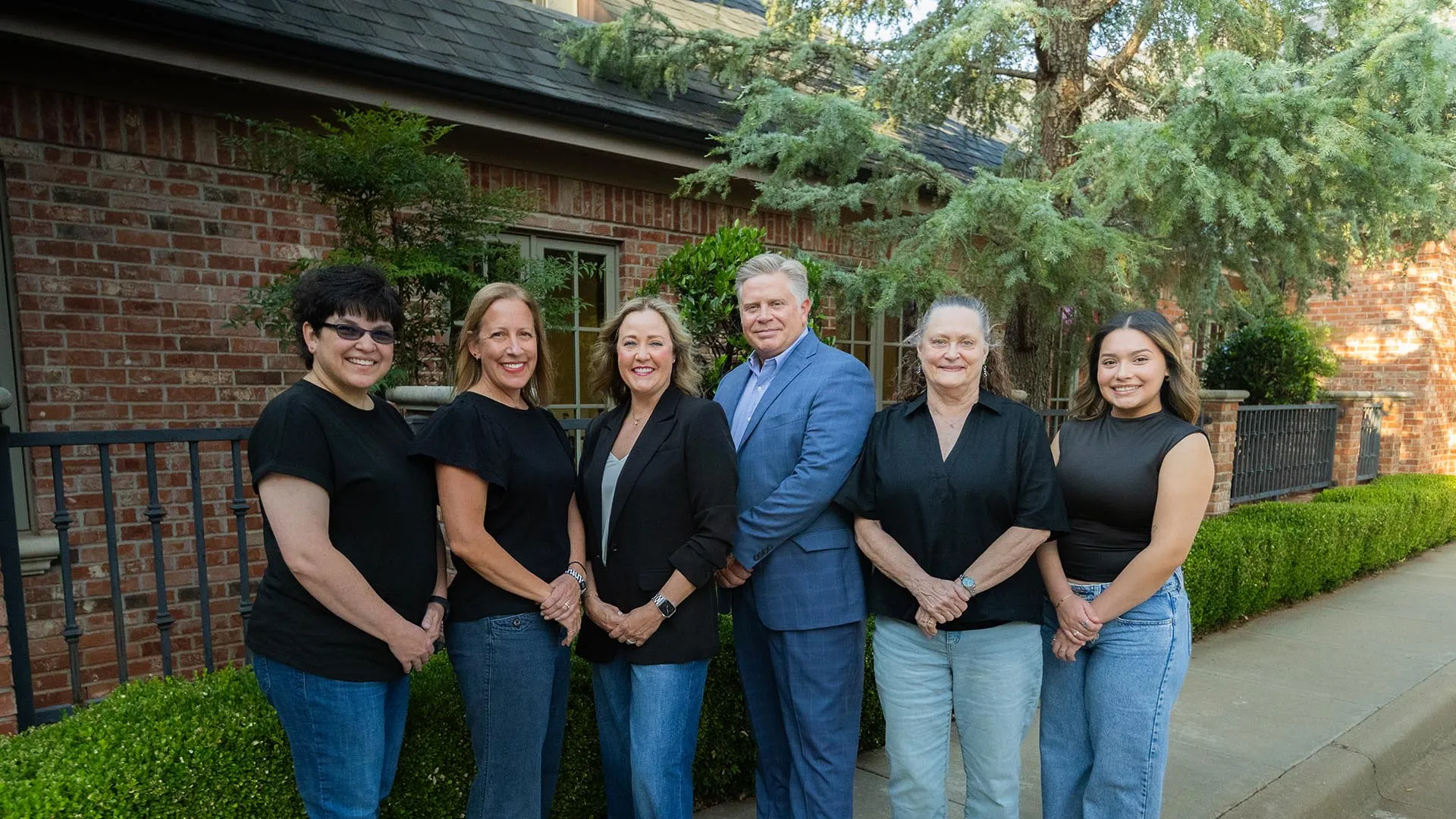 Group of six diverse professionals standing outdoors near brick building and greenery, smiling at the camera.
