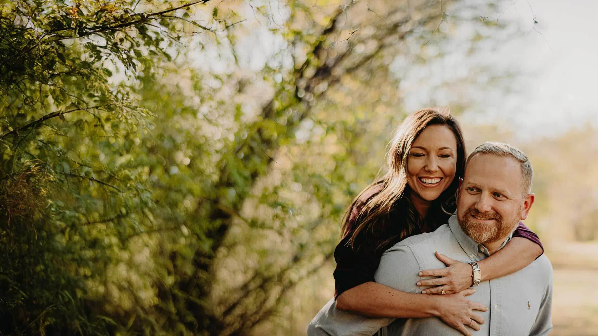 Happy couple enjoying a piggyback ride outdoors surrounded by lush green trees and warm sunlight.