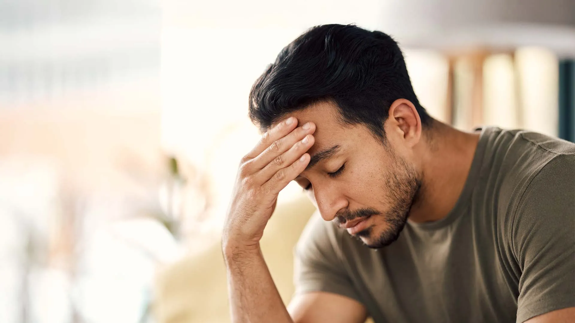 Man sitting with eyes closed and hand on forehead appearing stressed or deep in thought indoors.