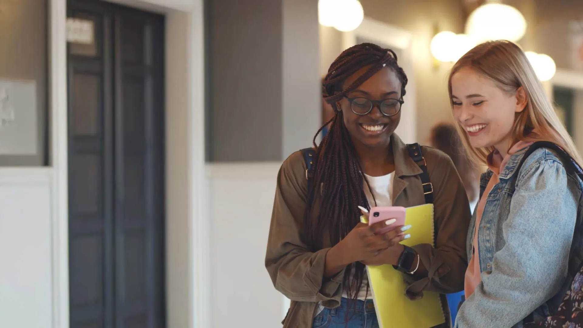Two smiling female students looking at a smartphone in a school hallway with backpacks and notebooks.
