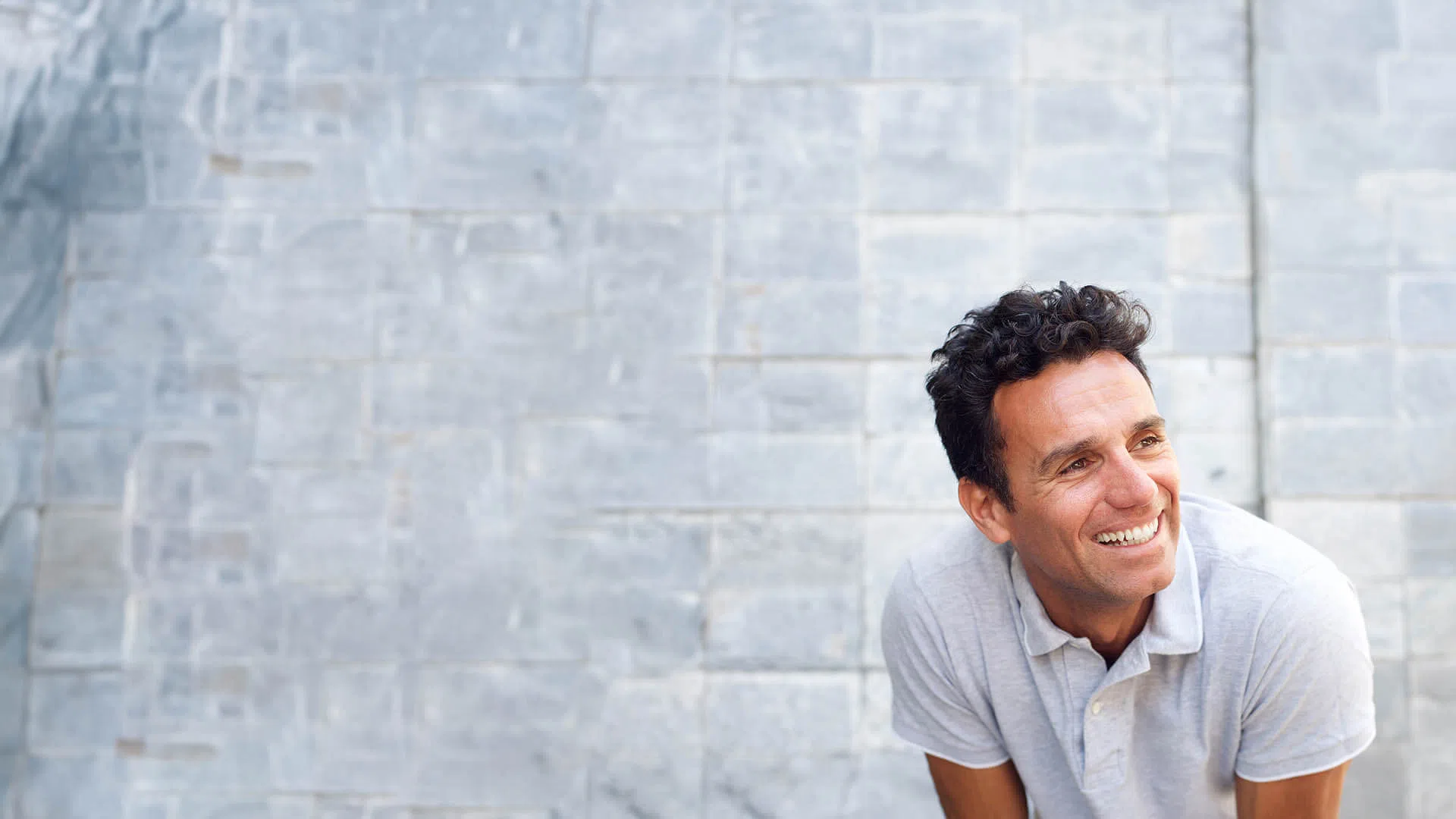 Smiling man with curly hair wearing a light grey polo shirt against a gray stone wall background.