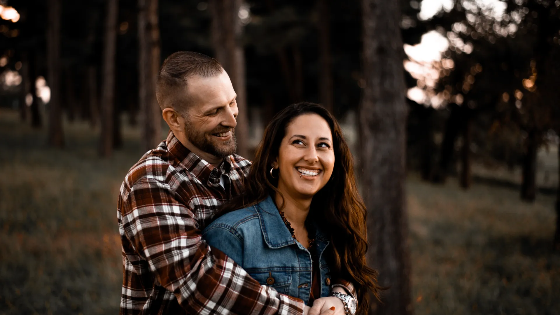 Smiling couple embracing in a forest during golden hour, showcasing warmth and affection in nature.