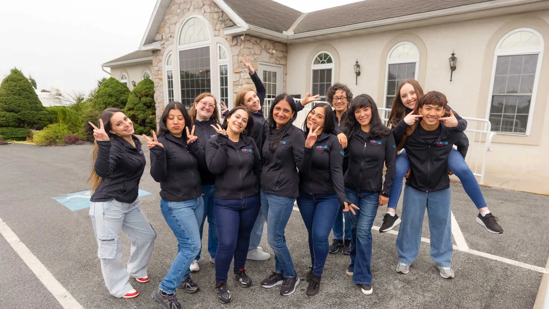 Group of diverse young adults wearing matching jackets posing cheerfully outside a stone and stucco building.