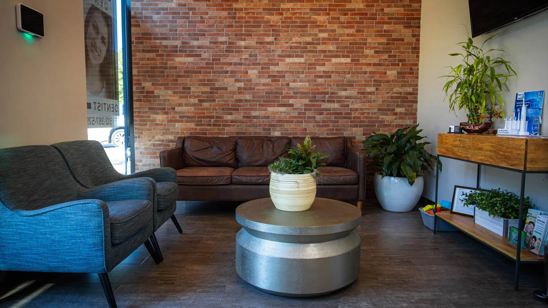 Modern waiting room with leather sofa, blue chairs, round metal table, brick wall, and green plants.