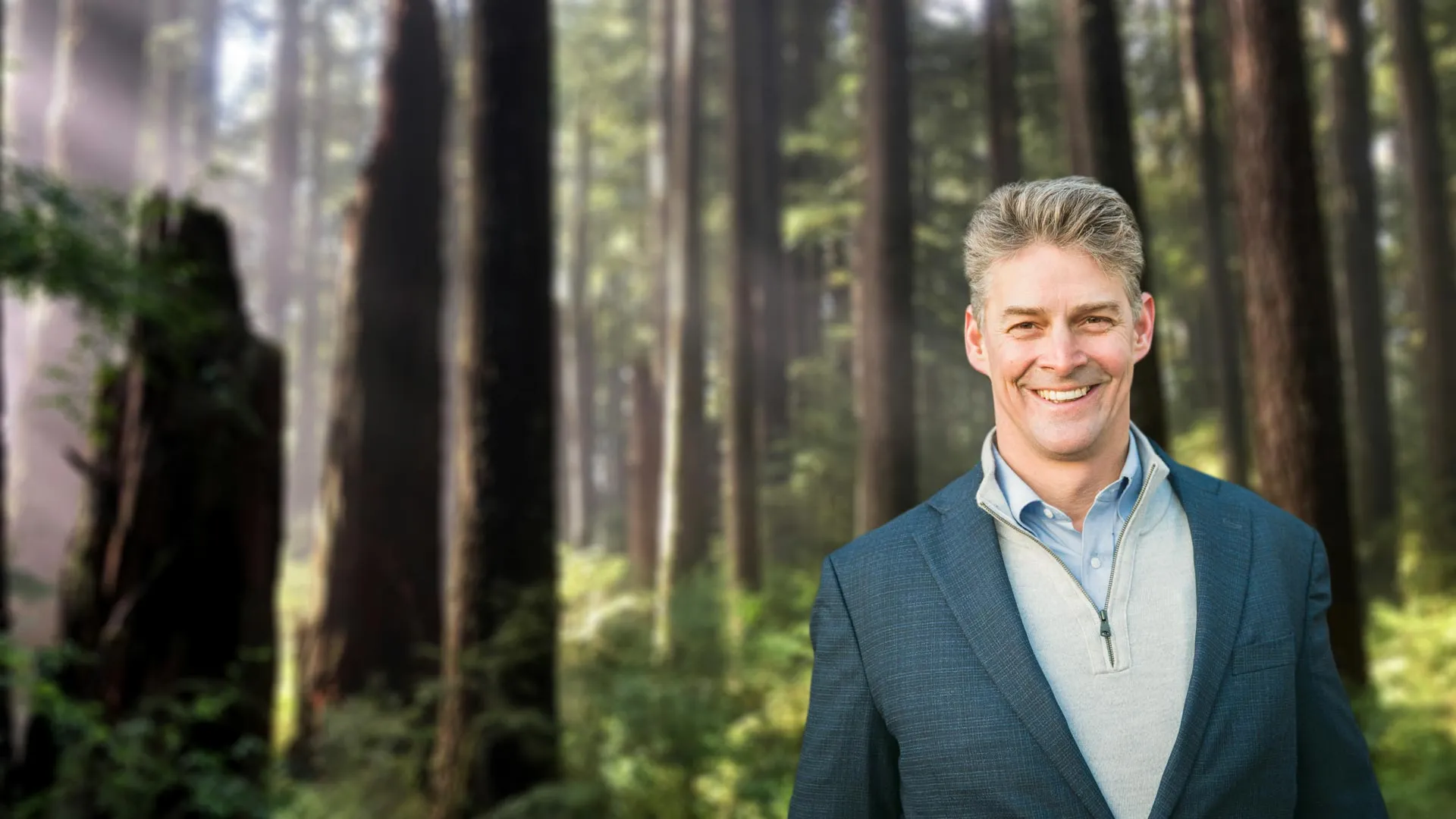 Smiling middle-aged man in blazer standing in a sunlit forest with tall trees and greenery in background