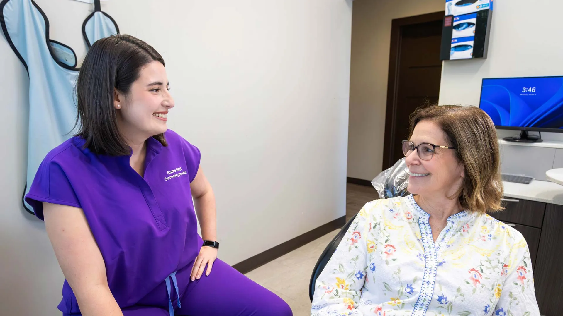 Dental hygienist in purple scrubs smiling and talking with a female patient in a dental office