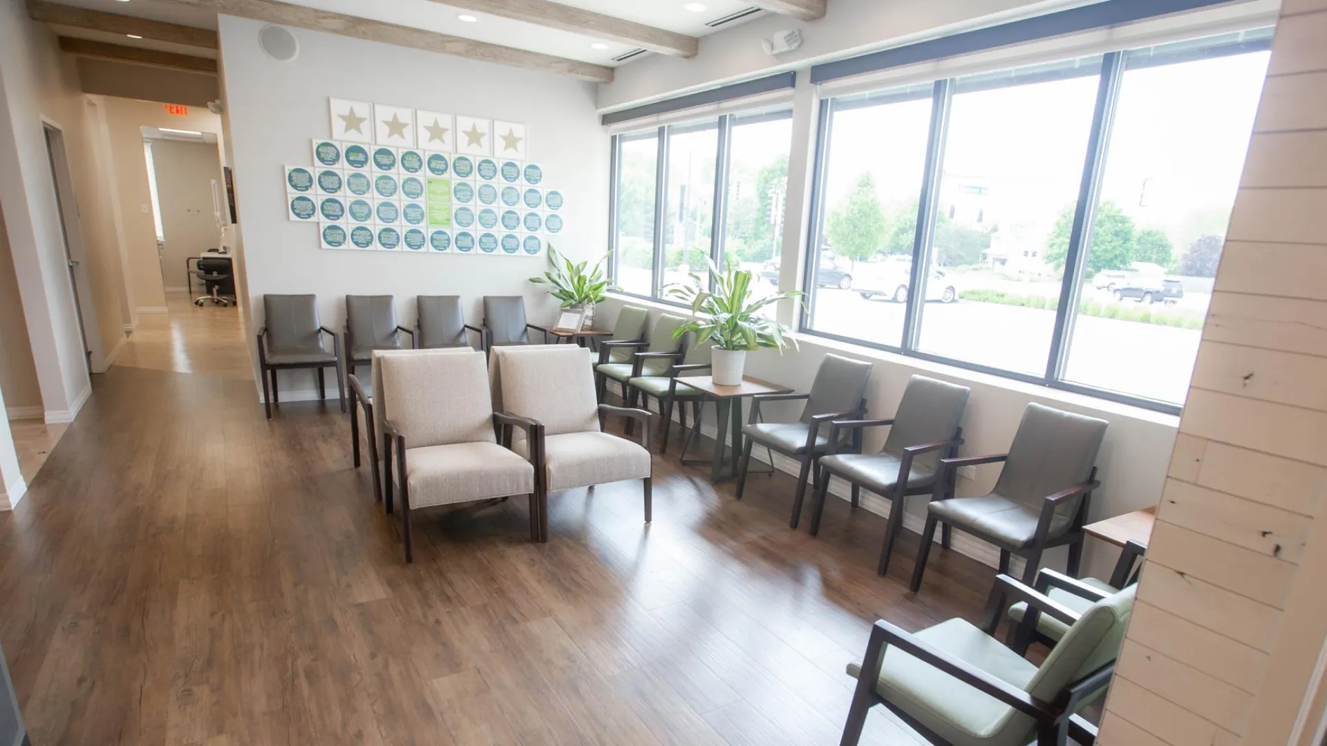Bright waiting room with rows of chairs, large windows, plants, and wooden flooring in a medical or office setting.