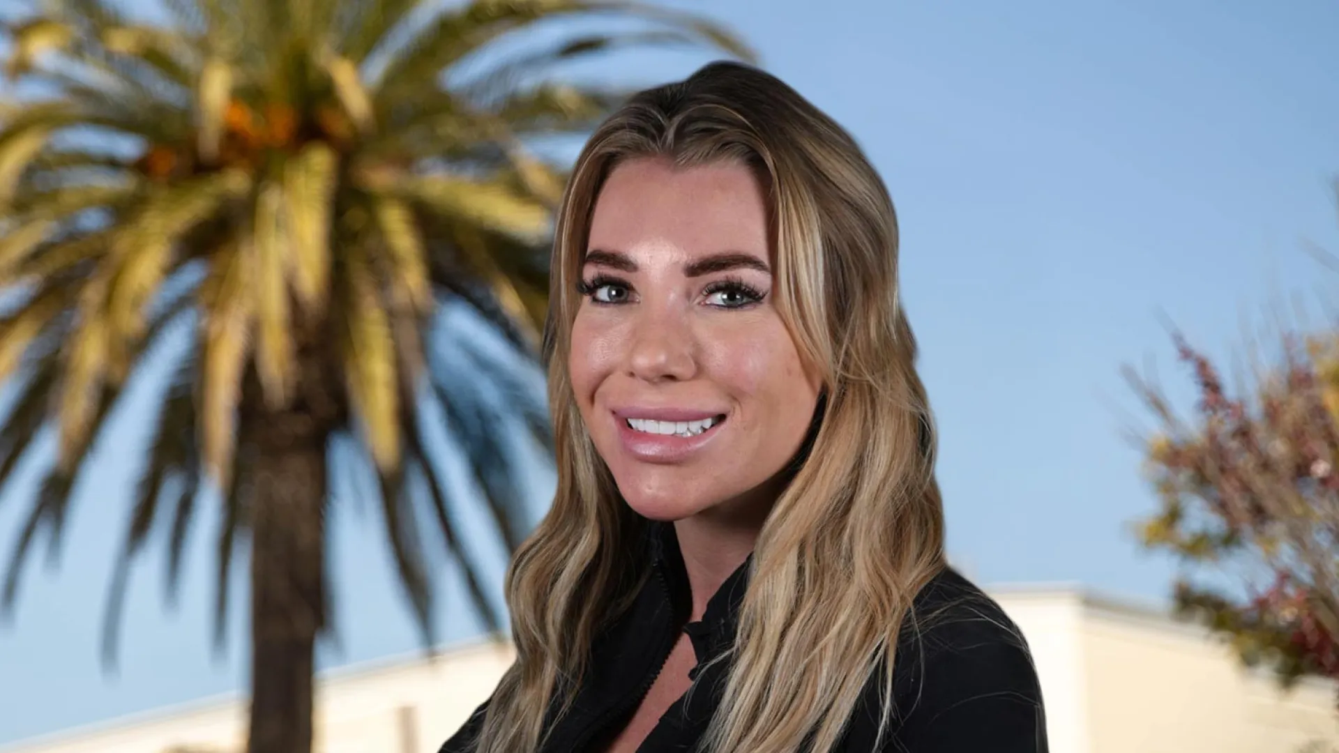 Smiling woman with long hair and palm trees in the background under a clear sky.