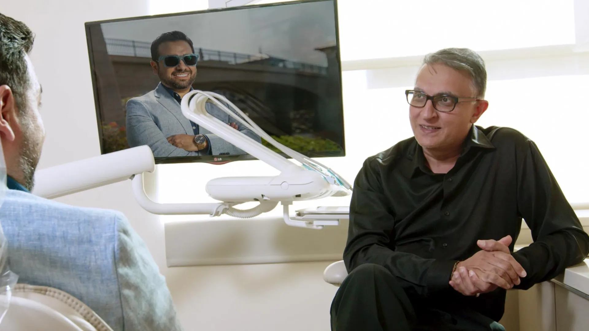 A dentist consults with a patient while a video is displayed on a screen in the background.