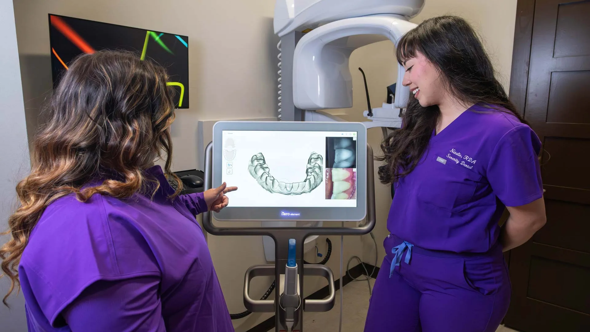 Two dental professionals in purple scrubs review 3D dental scan images on a monitor during consultation.