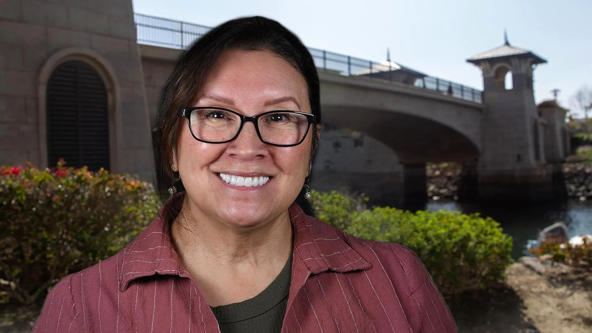 Smiling woman in front of a scenic bridge and water, surrounded by lush greenery and flowers.
