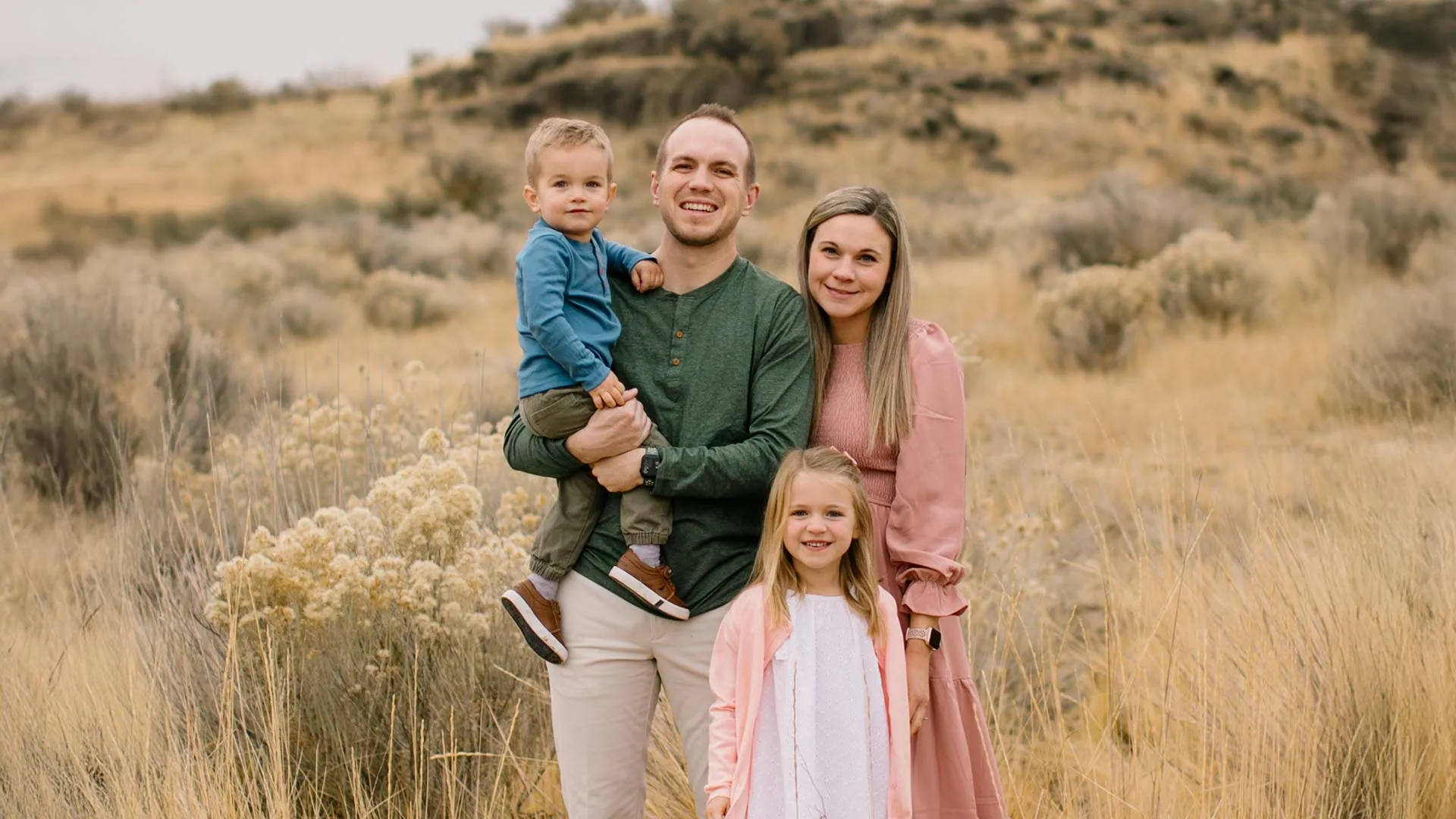 Smiling family of four posing in a grassy field with hills in the background on an overcast day