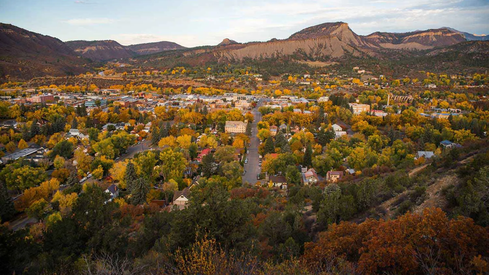 Aerial view of a town surrounded by autumn trees and mountains under a partly cloudy sky.