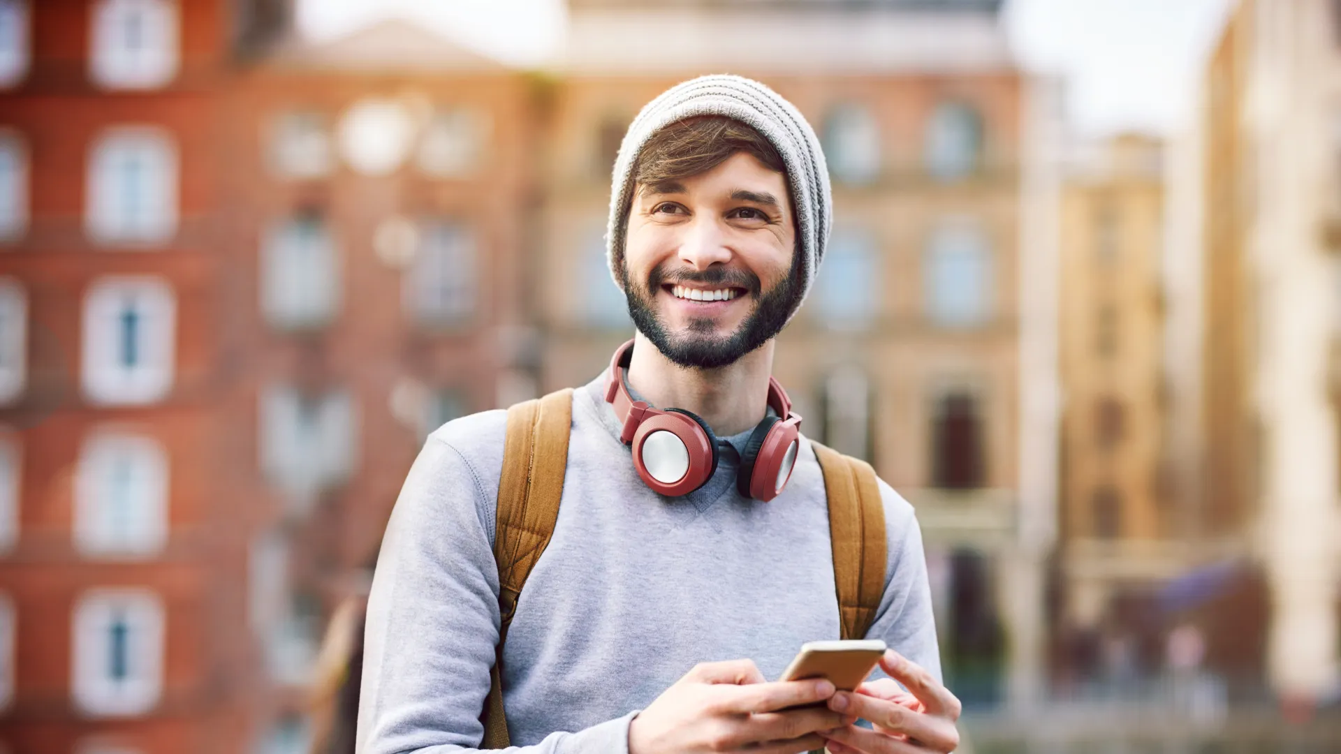 Young man wearing a beanie and headphones holds smartphone outdoors with urban buildings in background