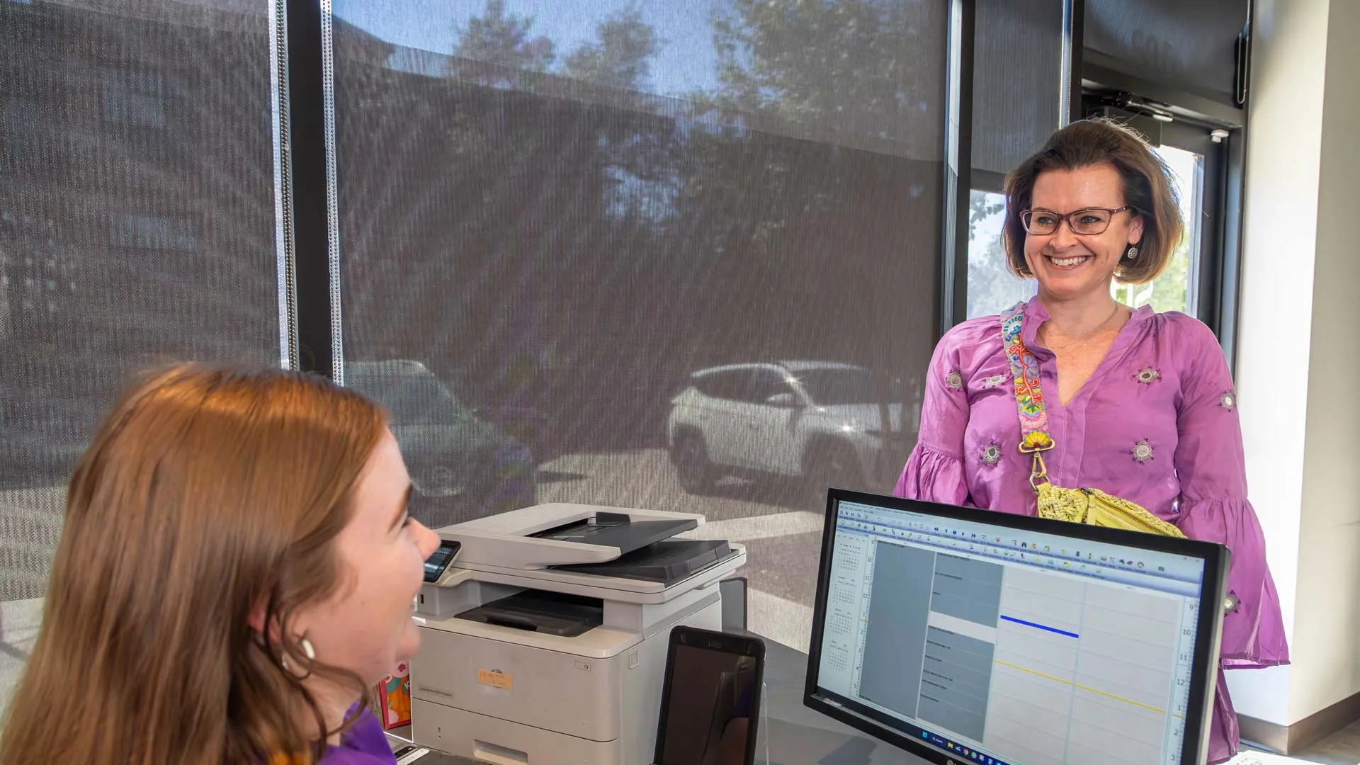 Receptionist in purple blouse smiling and assisting a woman with short hair at a modern office desk.