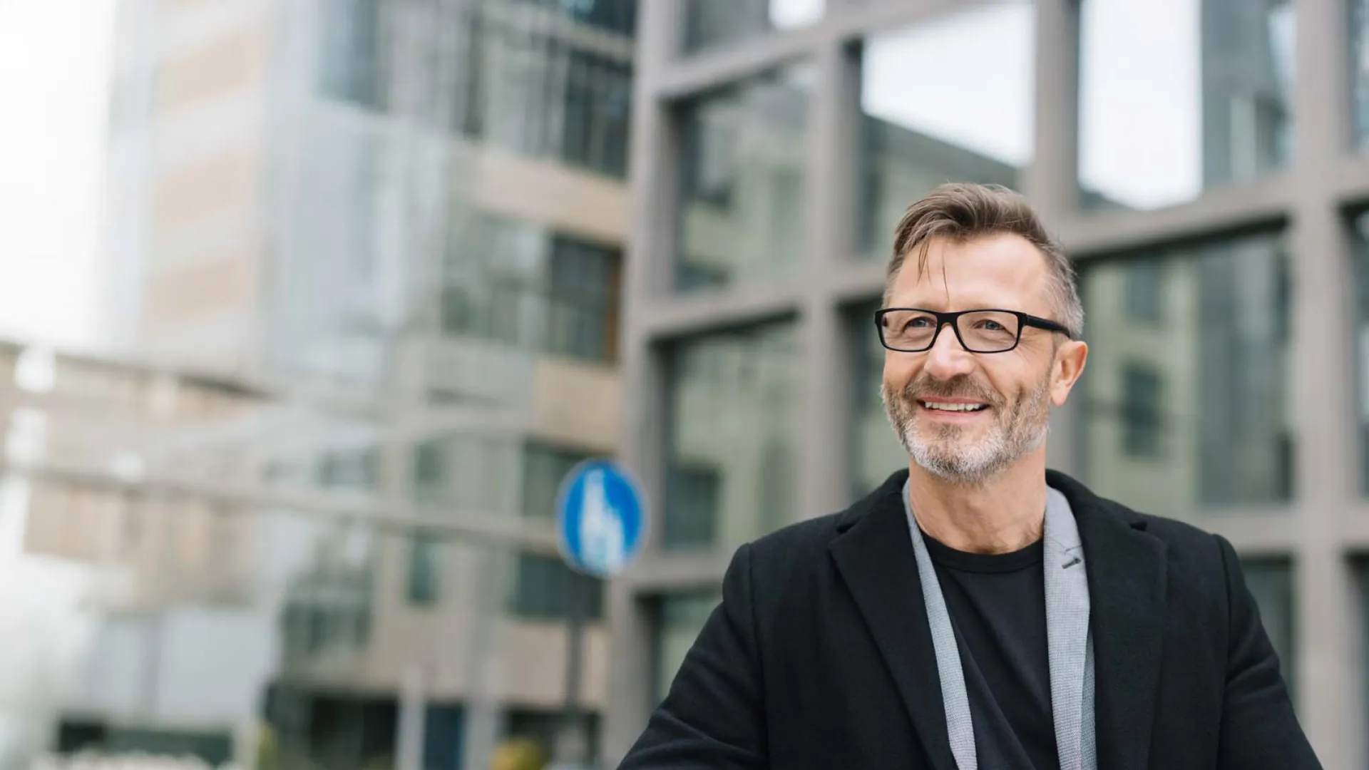 Smiling middle-aged man with glasses wearing a black jacket in an urban business district outdoors.