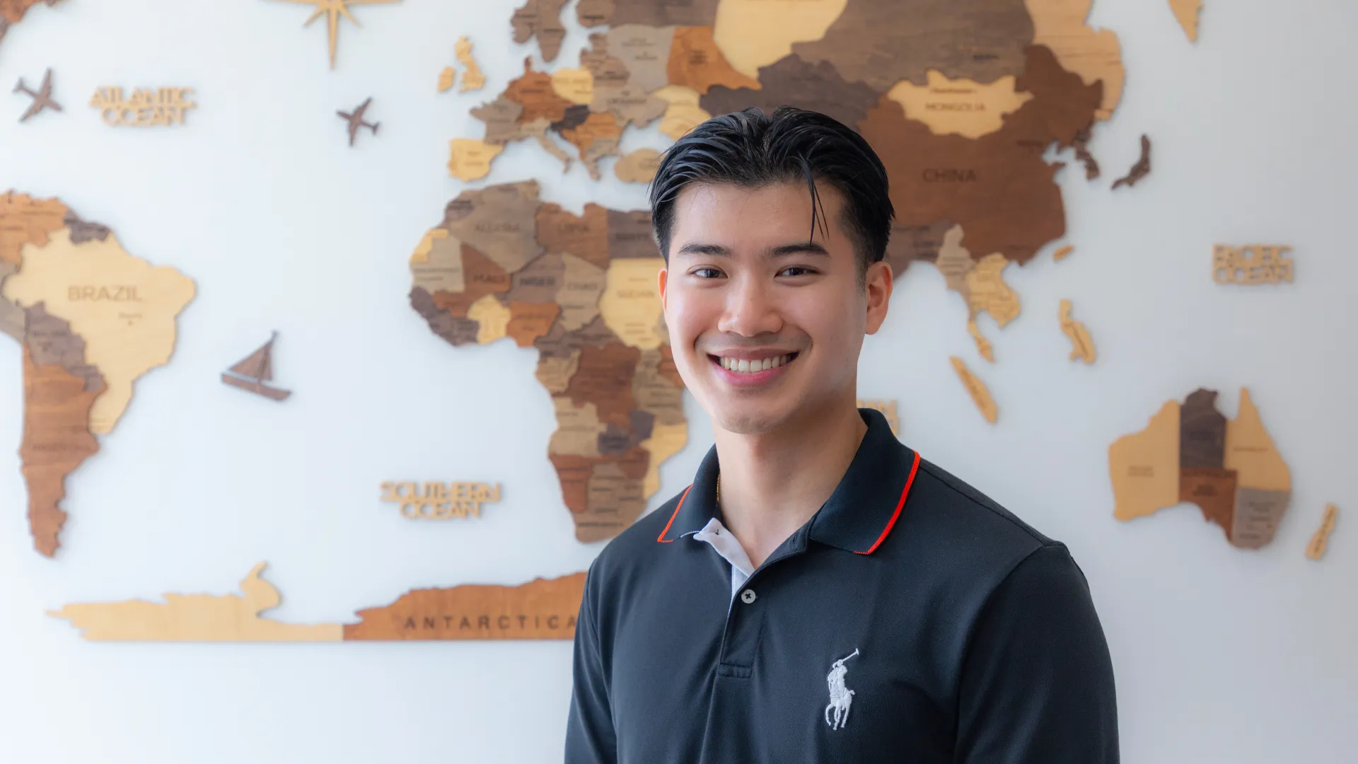 Smiling young man standing in front of a wooden world map wall decoration indoors.