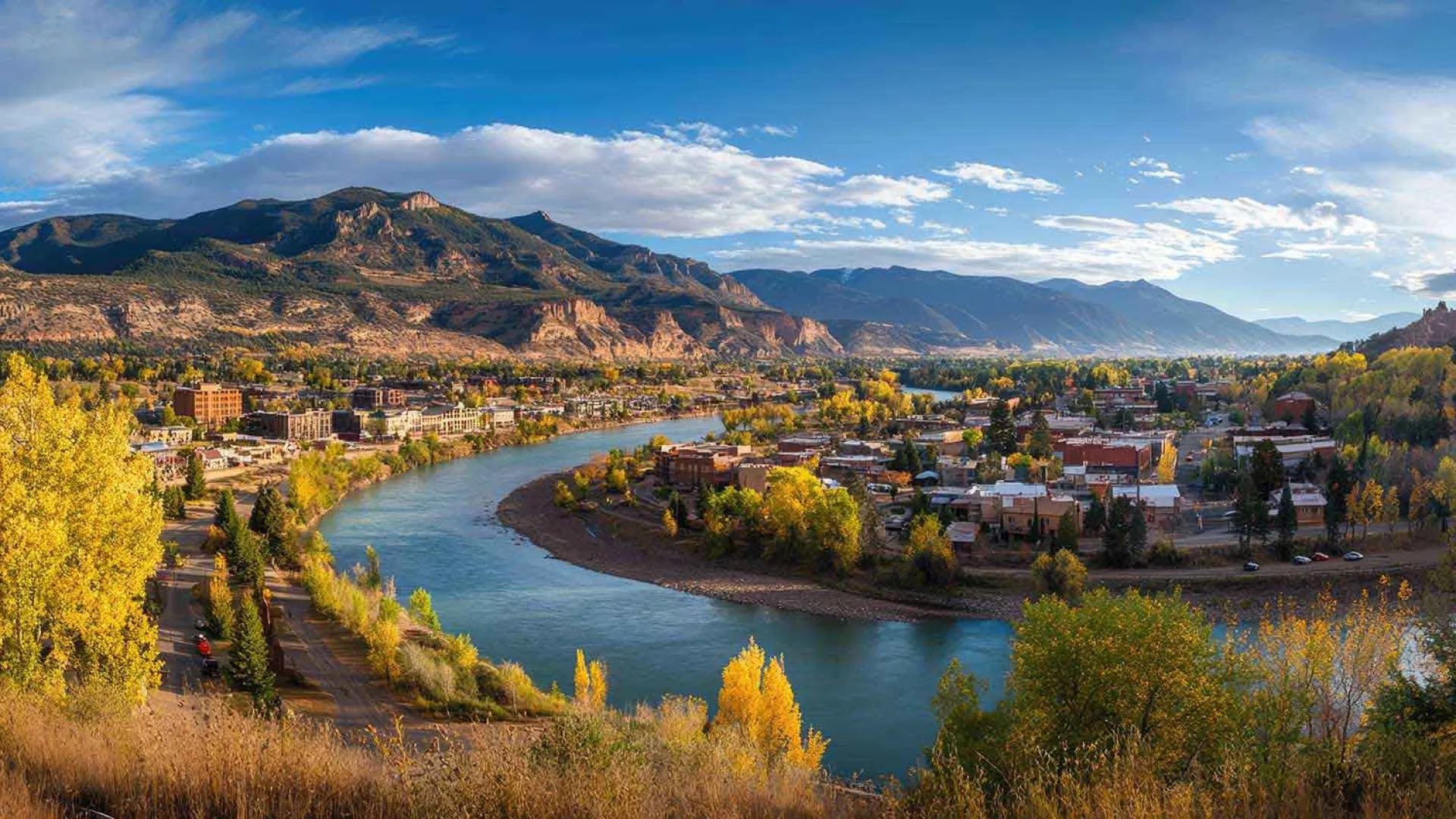 A river curves through a town with autumn trees and mountain ranges under a partly cloudy blue sky.