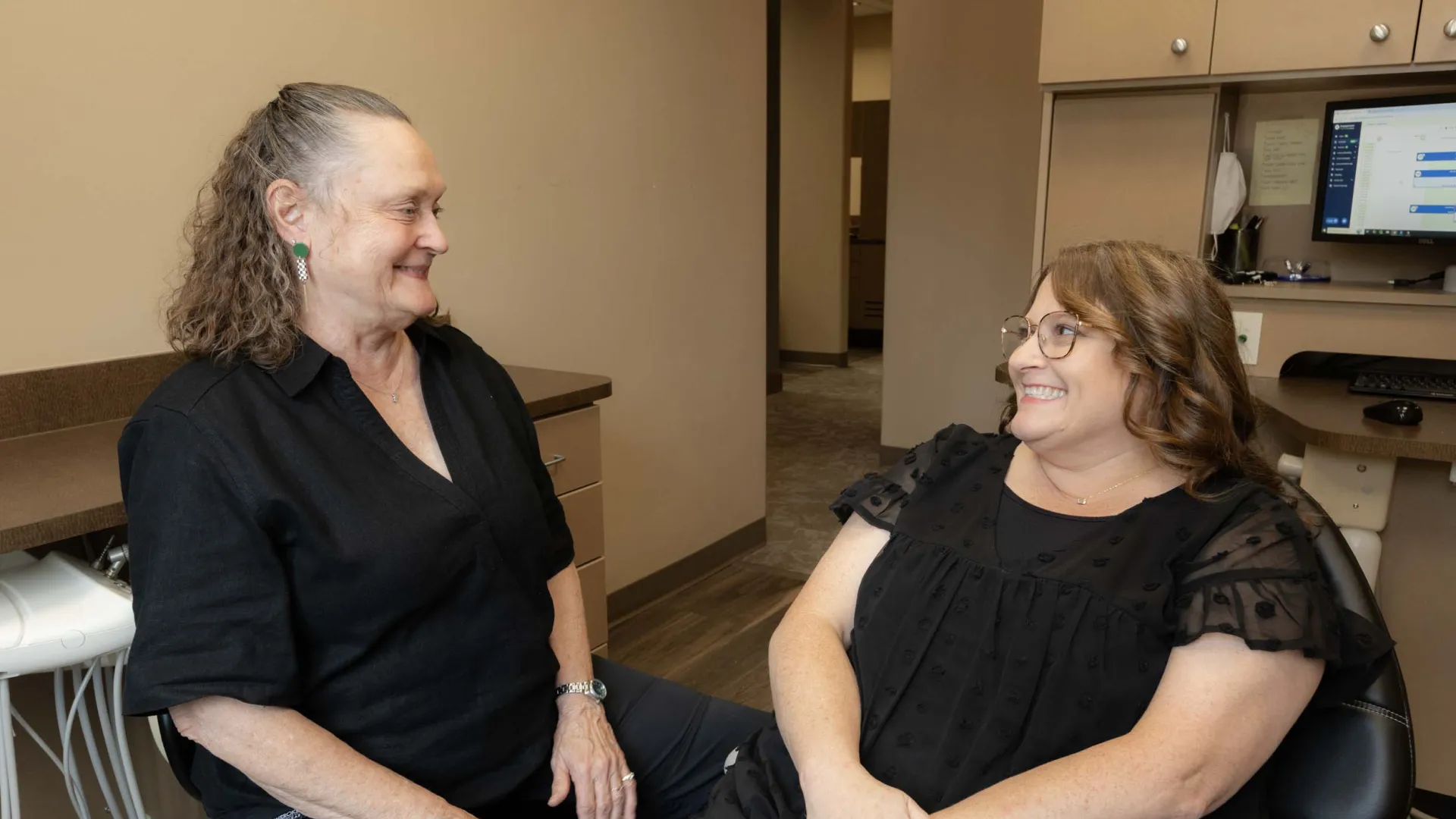 Two women smiling and chatting in a modern office with beige walls and a computer workstation.