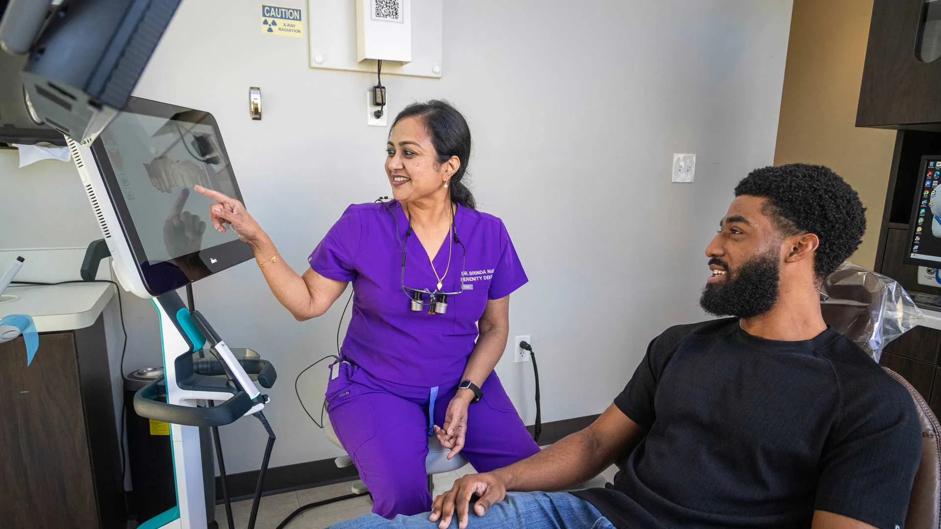 Dentist in purple scrubs showing dental x-rays on screen to male patient in clinic chair.