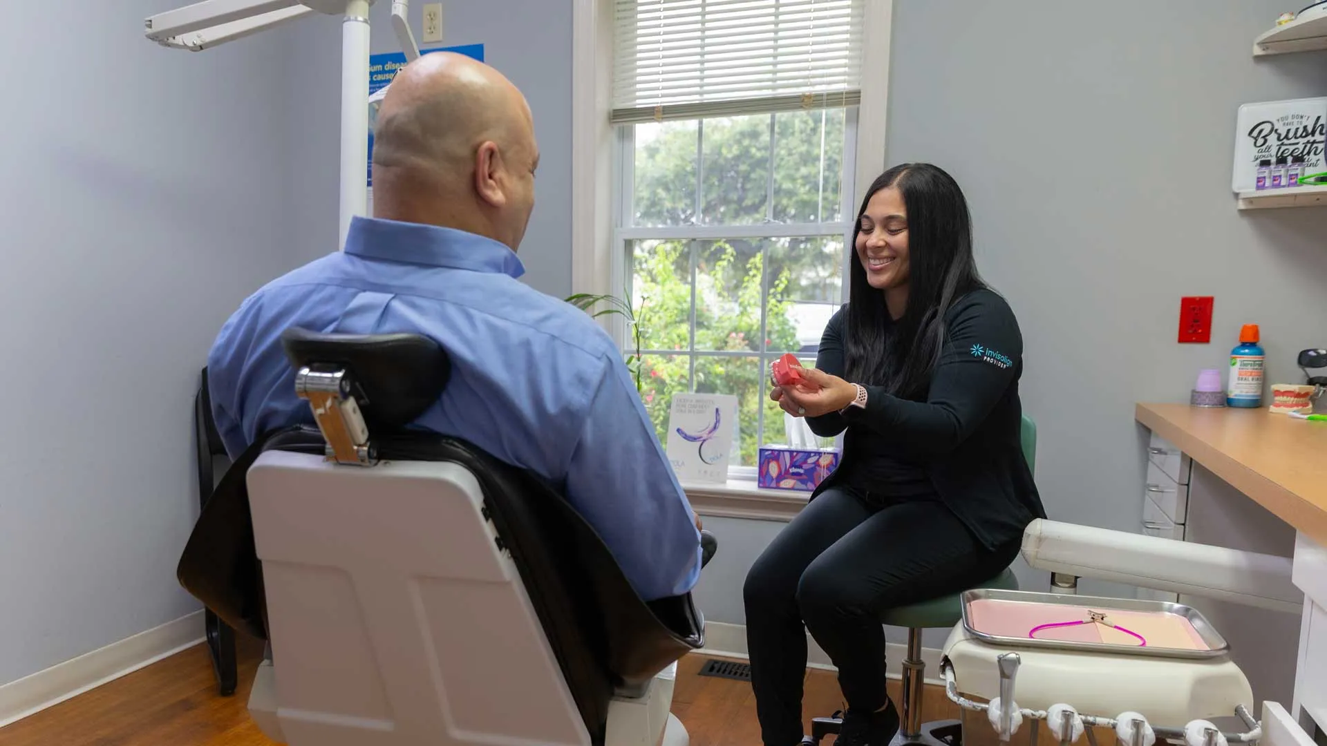Dental professional explaining teeth aligner to male patient in modern clinic with large window lighting.