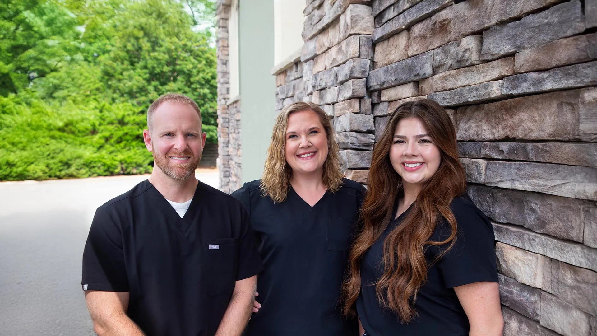 Three healthcare professionals in black scrubs smiling outside next to a stone wall and green foliage.
