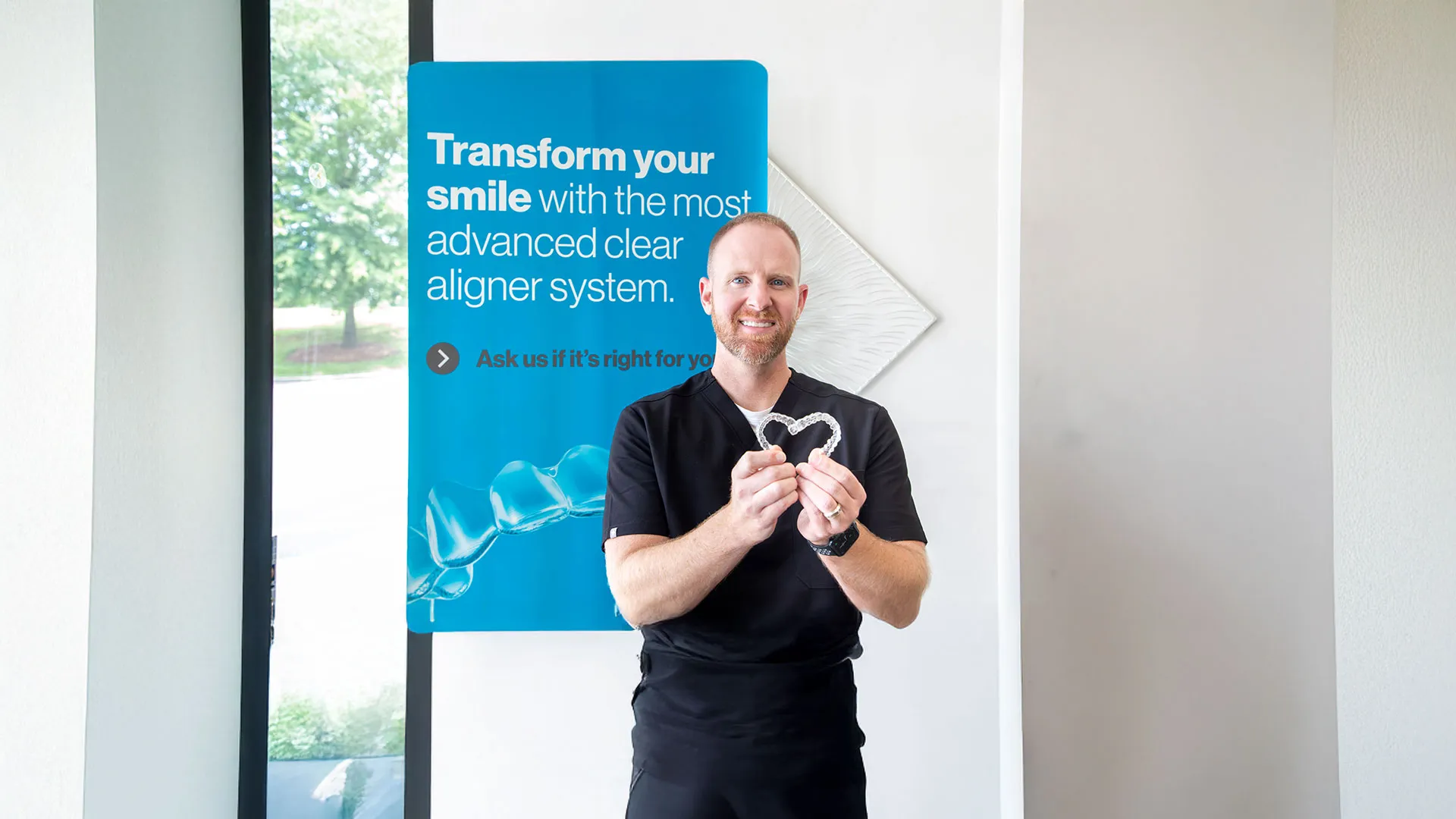 Smiling woman holding clear dental aligners in front of a promotional orthodontic banner about transforming your smile.