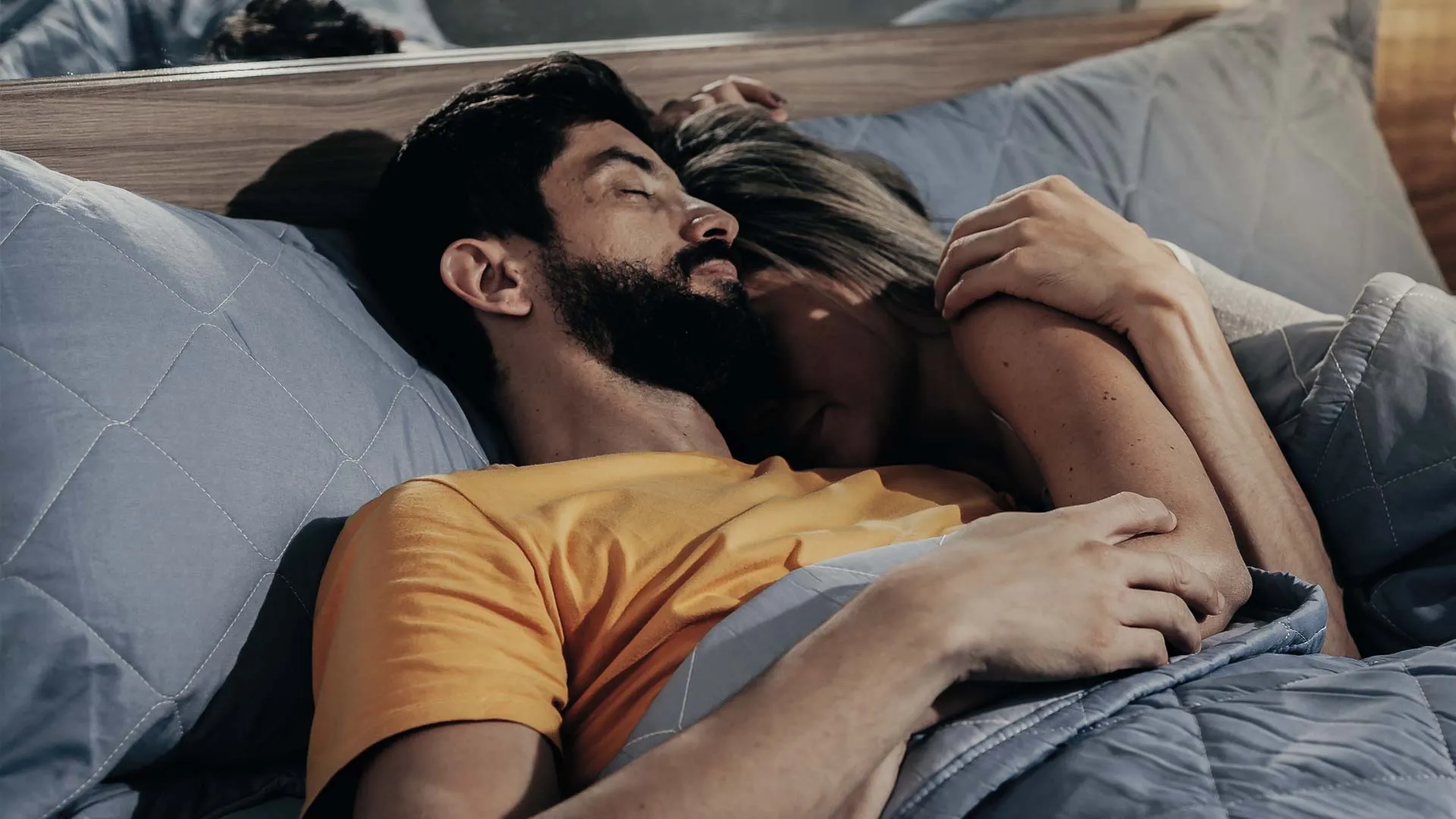 Couple cuddling closely in bed under a gray quilt, showing affection and comfort in a cozy setting.