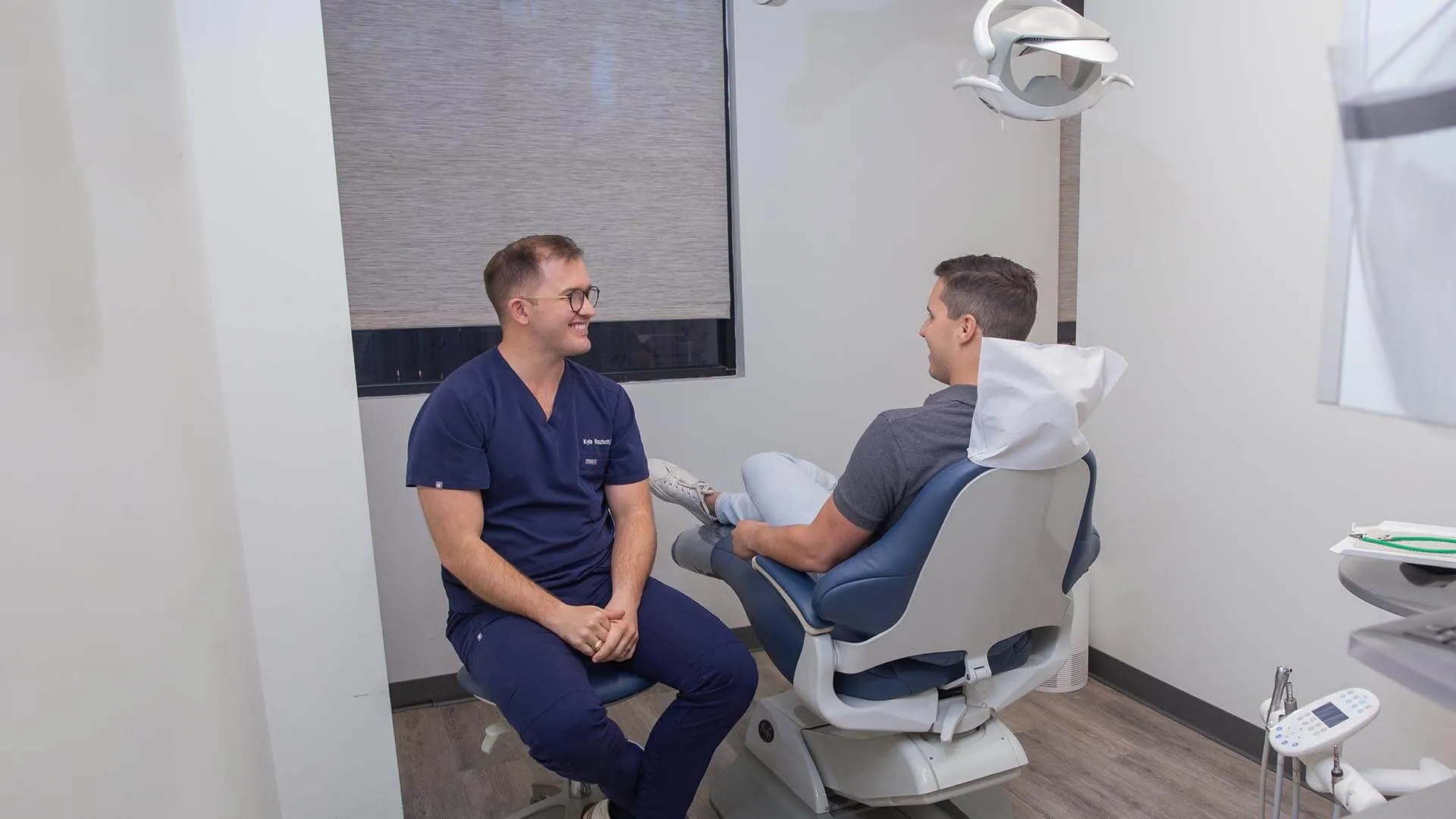 Dentist in navy scrubs consulting a male patient seated in a dental chair in a modern clinic room.