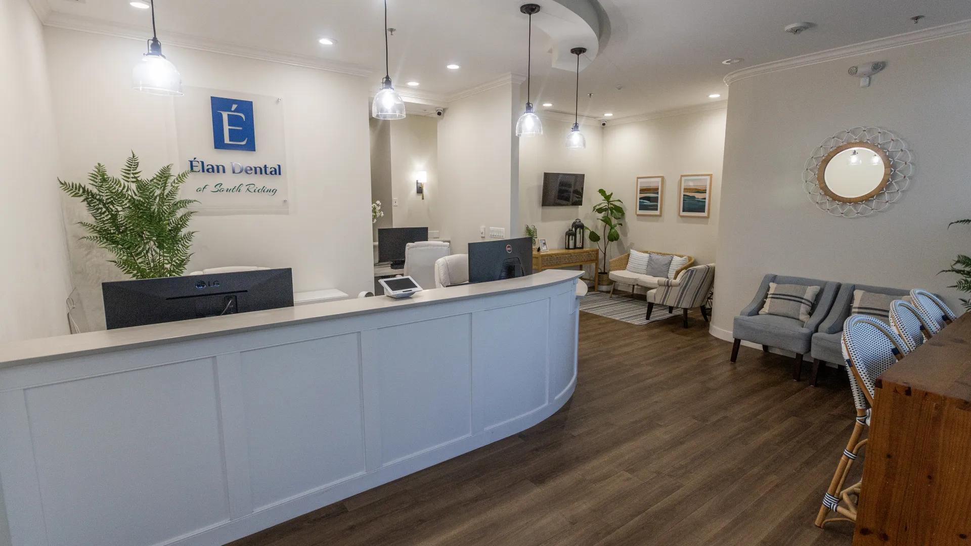 Modern dental office reception with white curved desk, comfortable seating, wood flooring, and pendant lighting.
