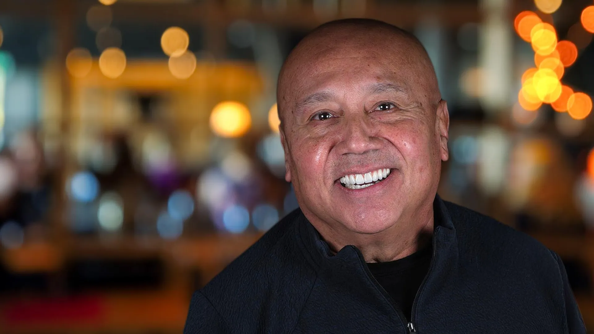 Smiling man with a warm expression, blurred lights in the background create a vibrant atmosphere.