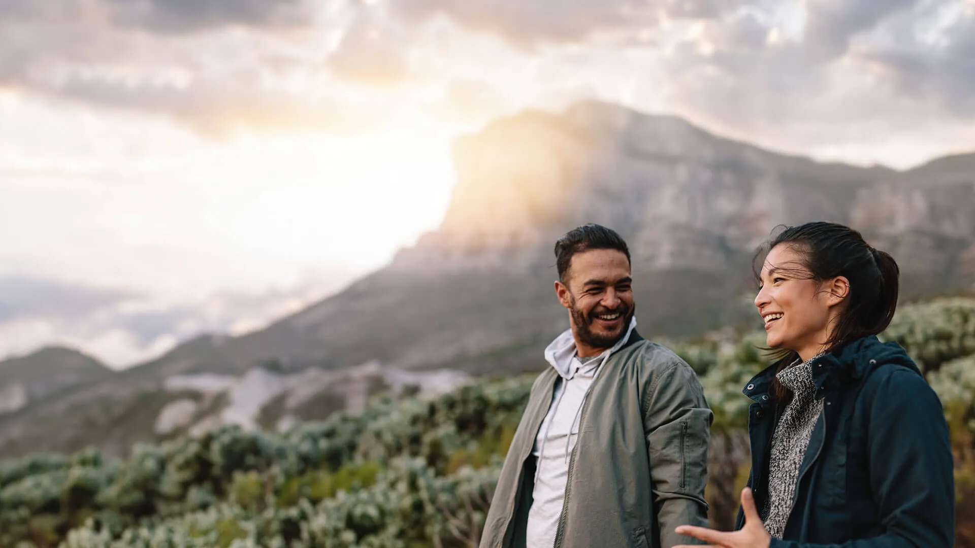 Smiling man and woman hiking outdoors with mountains and sunset in the background, enjoying nature and conversation.
