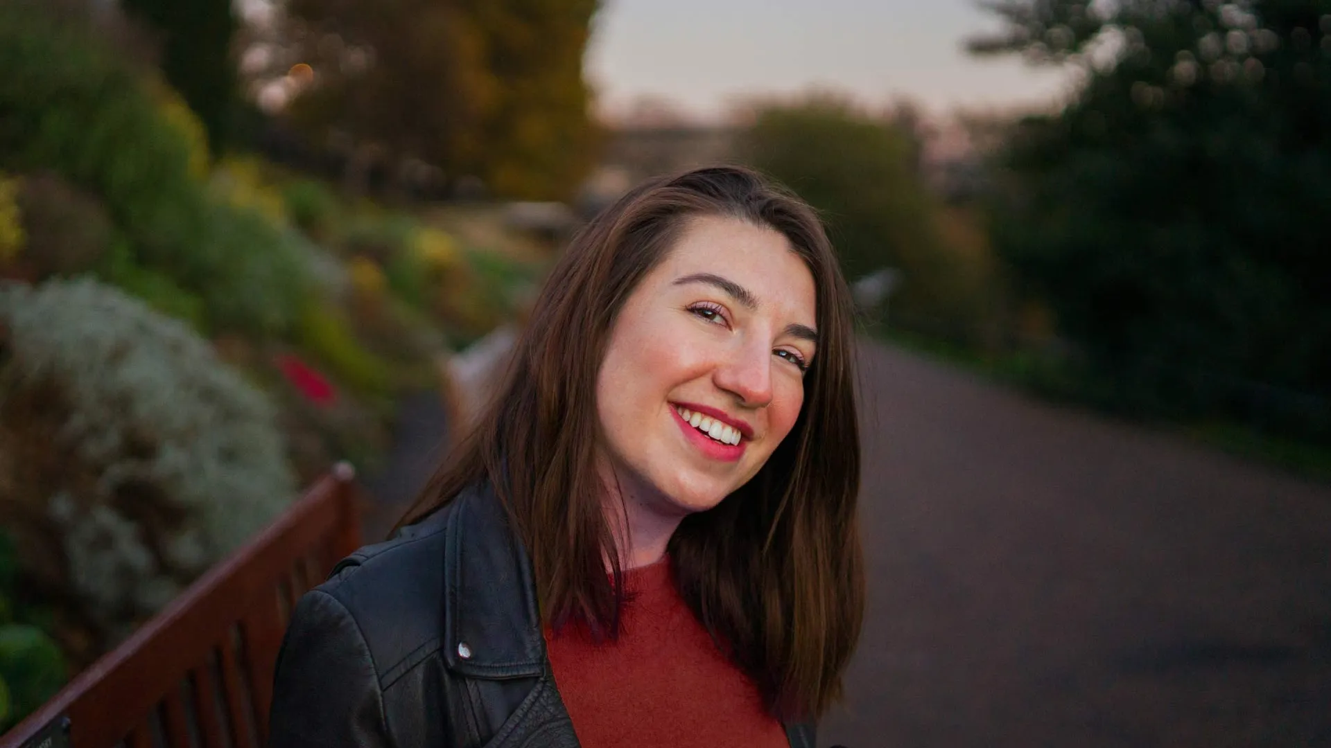 Smiling woman with brown hair wearing a black leather jacket and red sweater sitting on a bench in a park during sunset.
