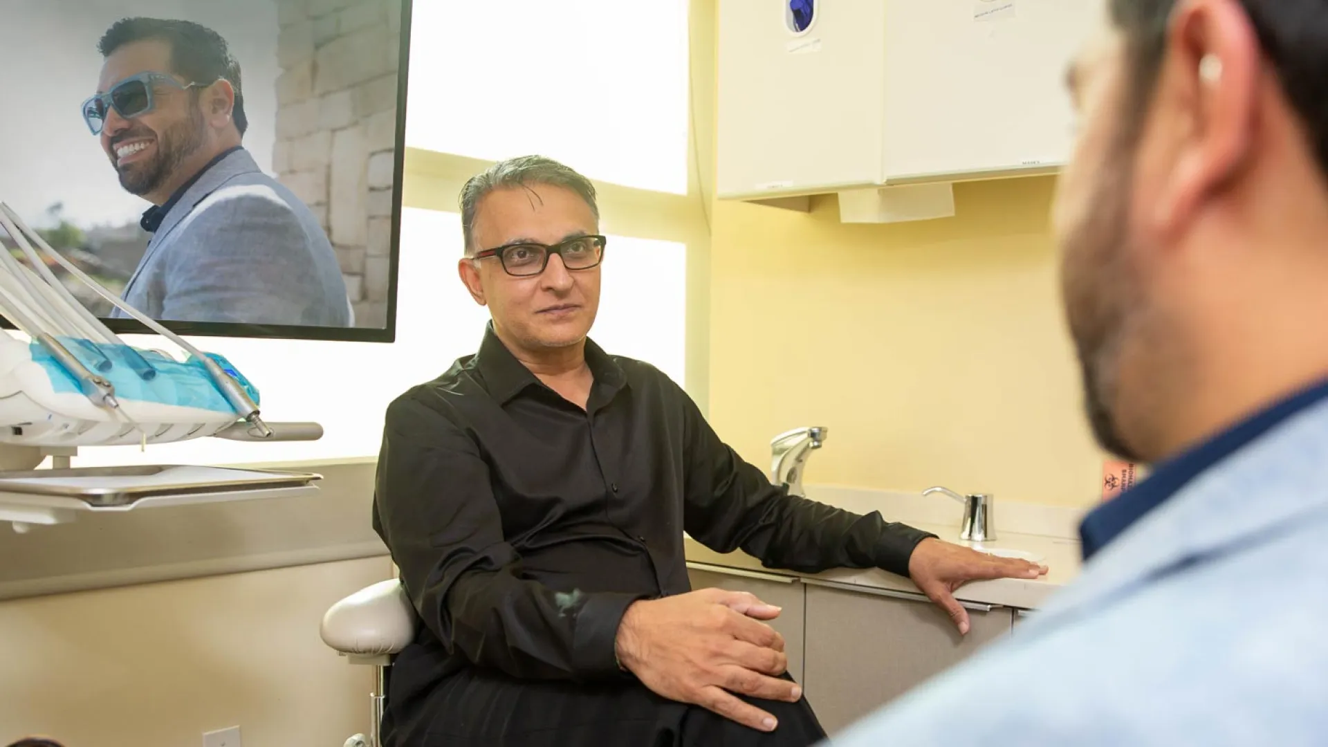 A dentist consulting with a patient in a well-lit clinic, featuring dental equipment and a monitor.