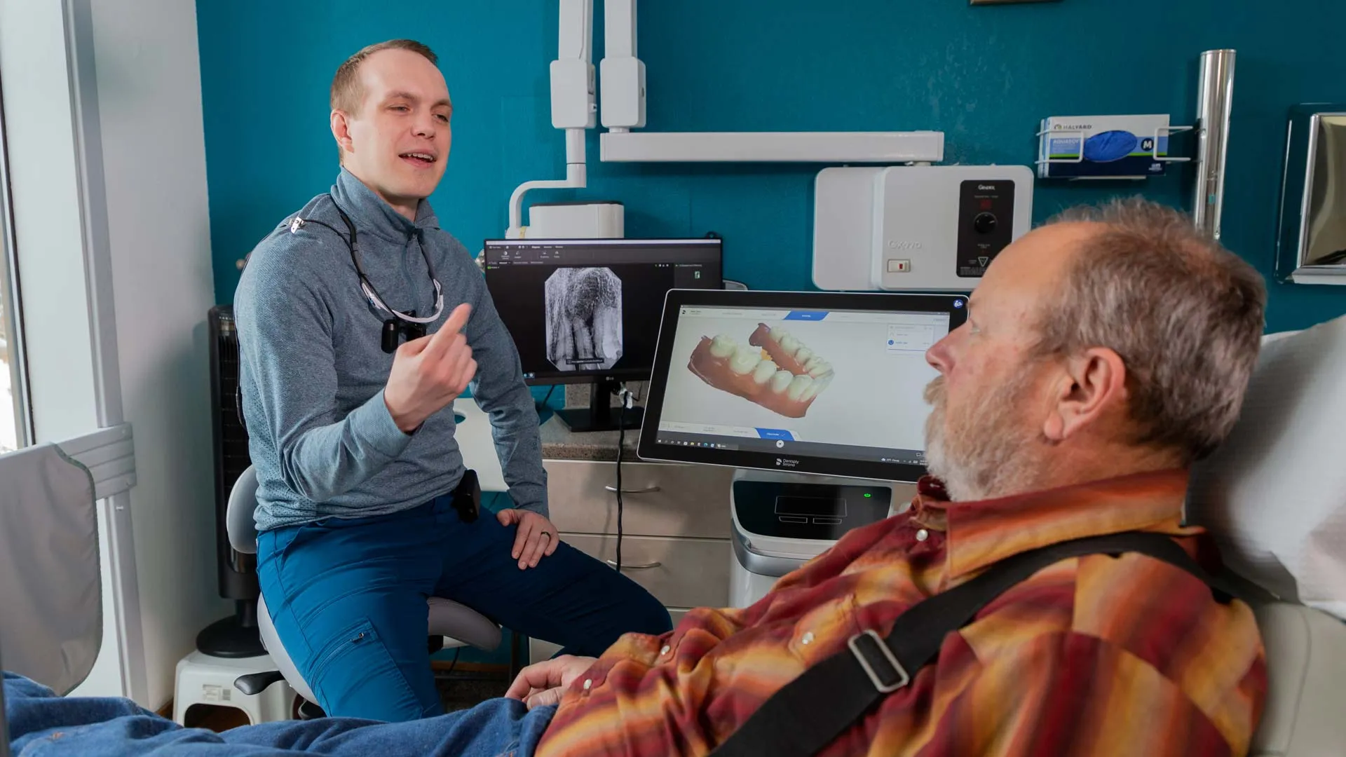 Dentist explaining dental treatment to elderly patient with 3D tooth scan and x-ray images on screens.