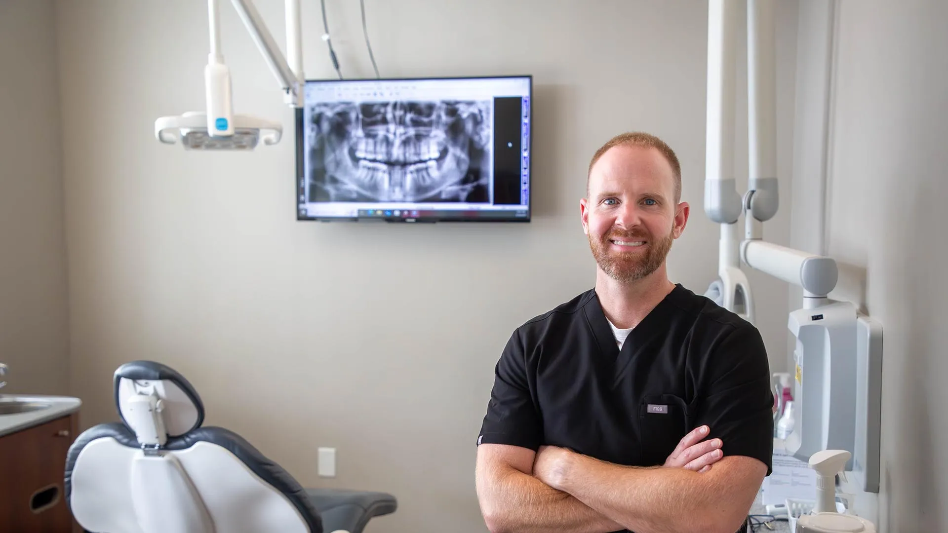 Smiling male dentist in black scrubs stands confidently in dental office with panoramic dental X-ray on screen