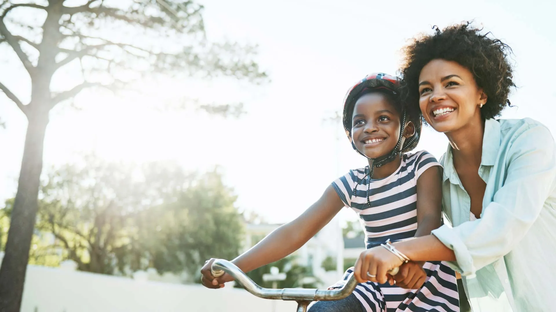 Mother and daughter smiling outdoors as the daughter learns to ride a bicycle wearing a helmet on a sunny day.