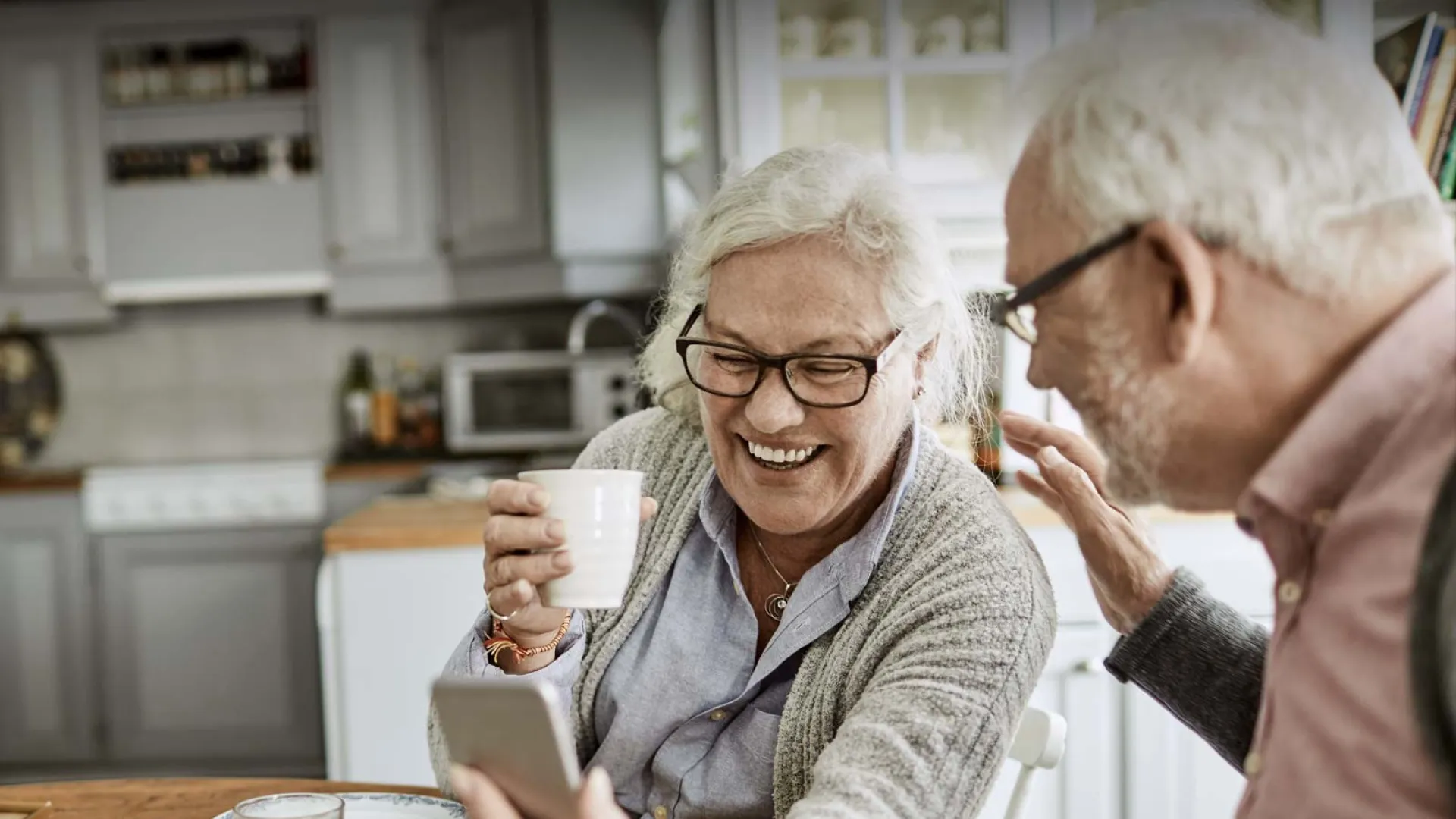 Happy seniors enjoying a moment together while looking at a smartphone in a cozy kitchen setting.