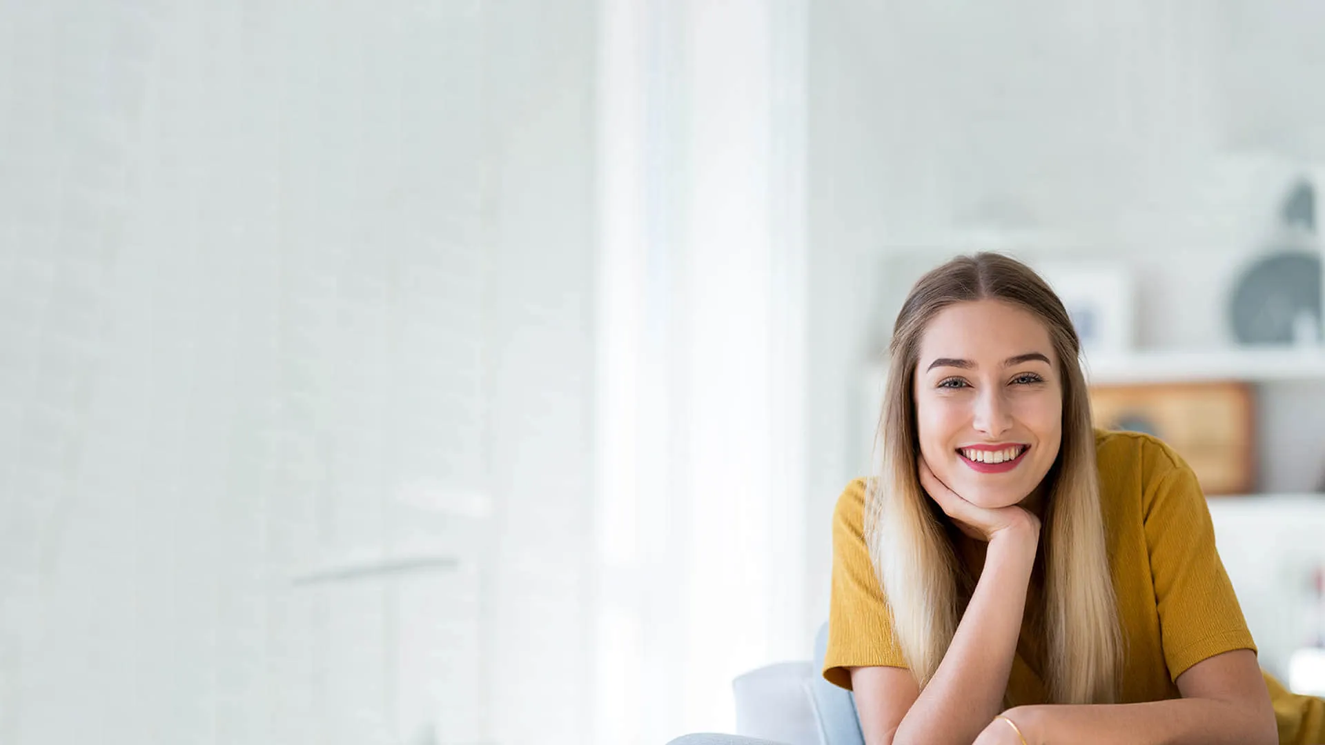 Smiling woman in yellow shirt sitting in bright, modern room with soft lighting and neutral decor.