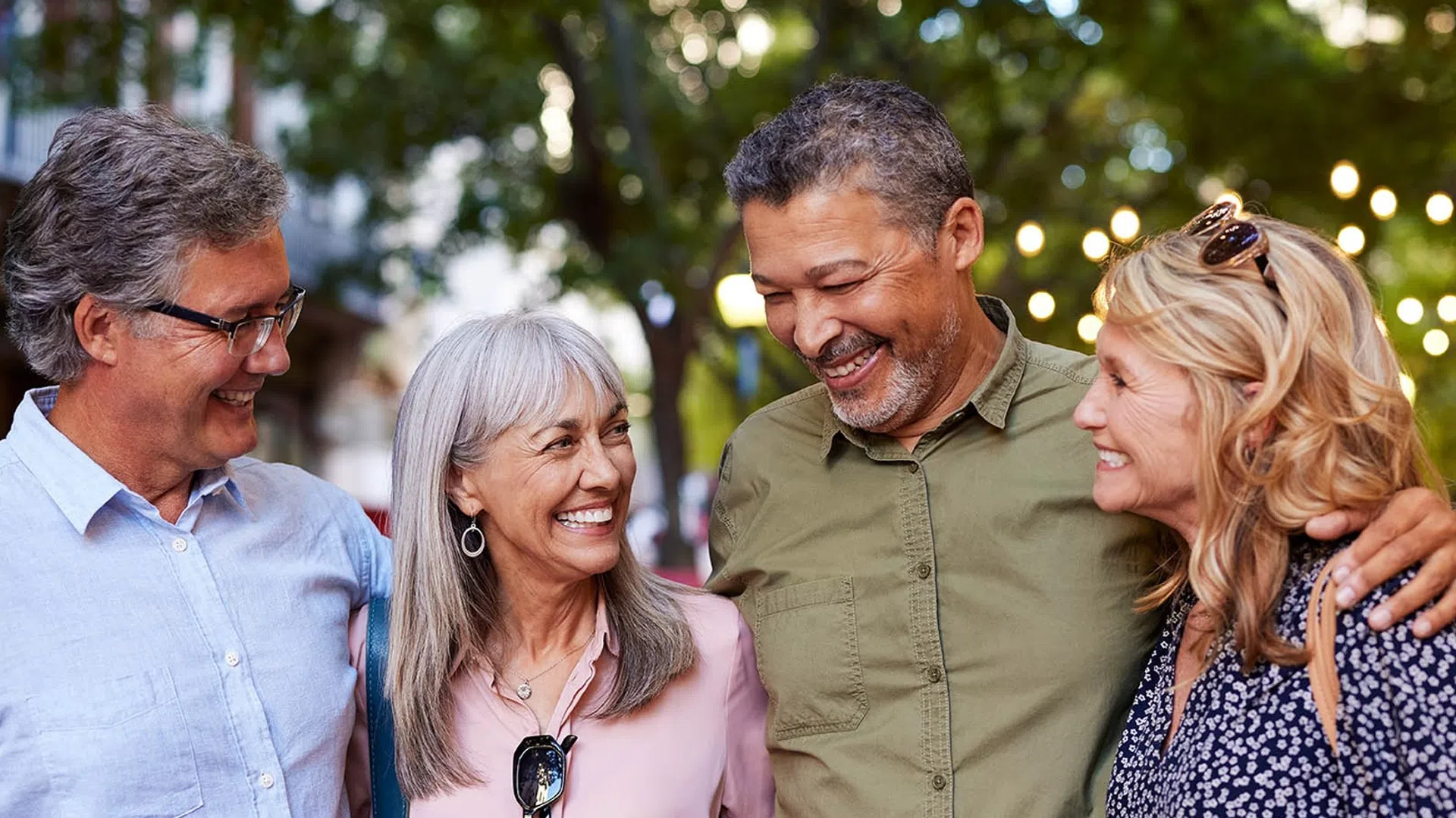 Four happy middle-aged friends smiling and chatting outdoors with trees and city lights in the background.
