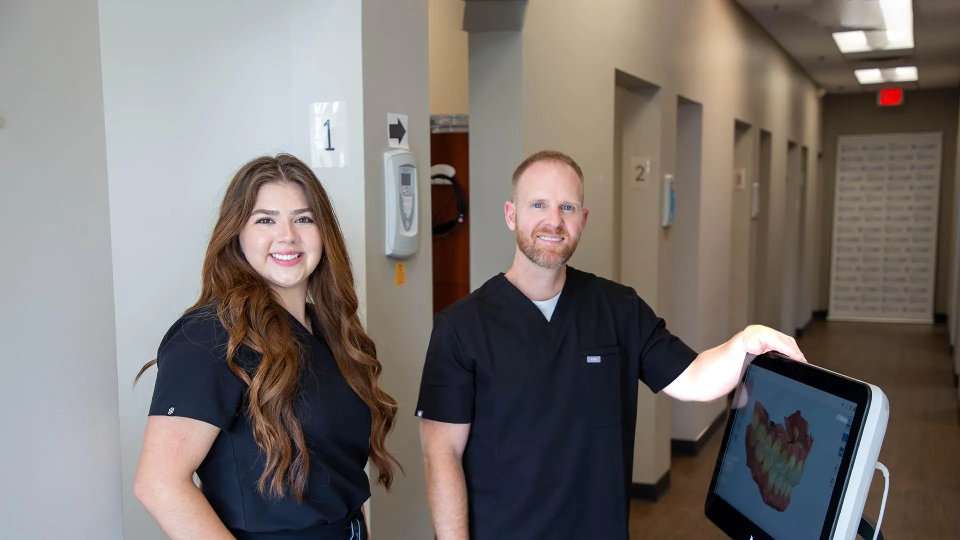 Two dental professionals in black scrubs stand in a clinic hallway with dental imaging equipment.