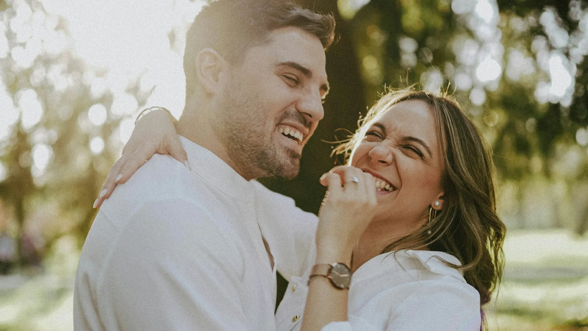 Happy couple in white shirts laughing and embracing outdoors with sunlight filtering through trees.