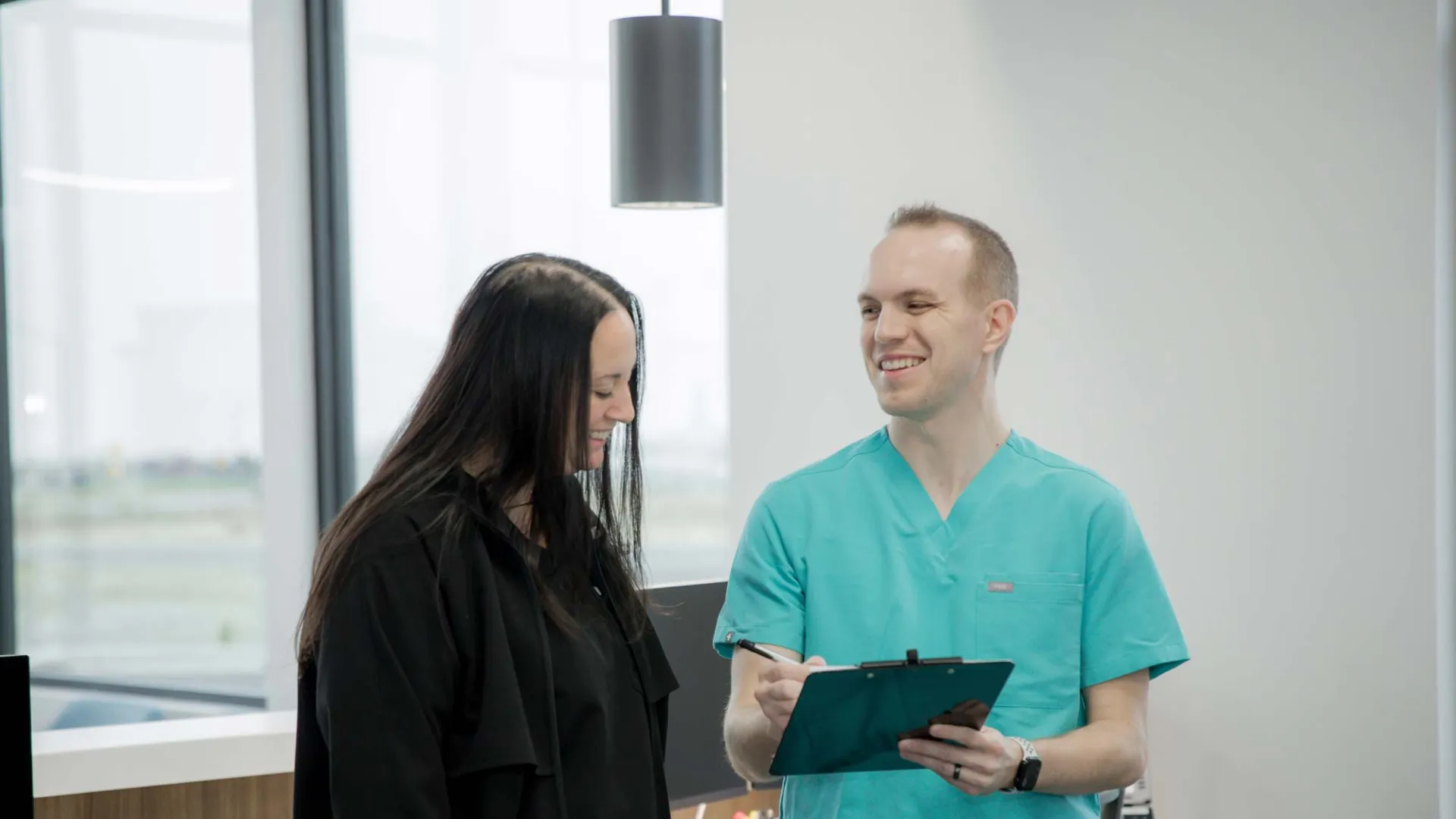 Healthcare professional in teal scrubs discussing information with woman in black in a bright modern office.