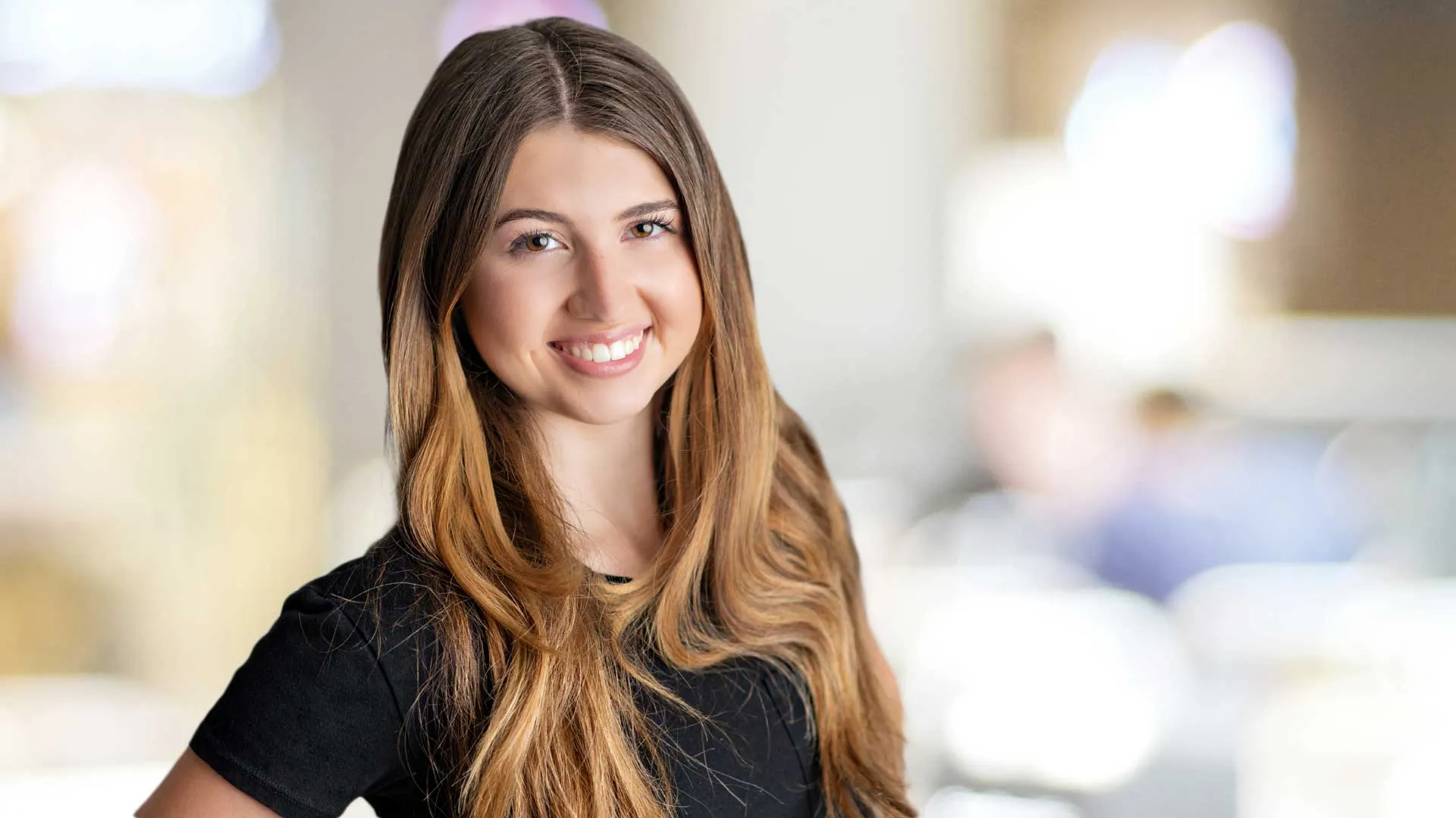 Smiling young woman with long brown hair wearing a black shirt, standing in a bright blurred indoor setting.