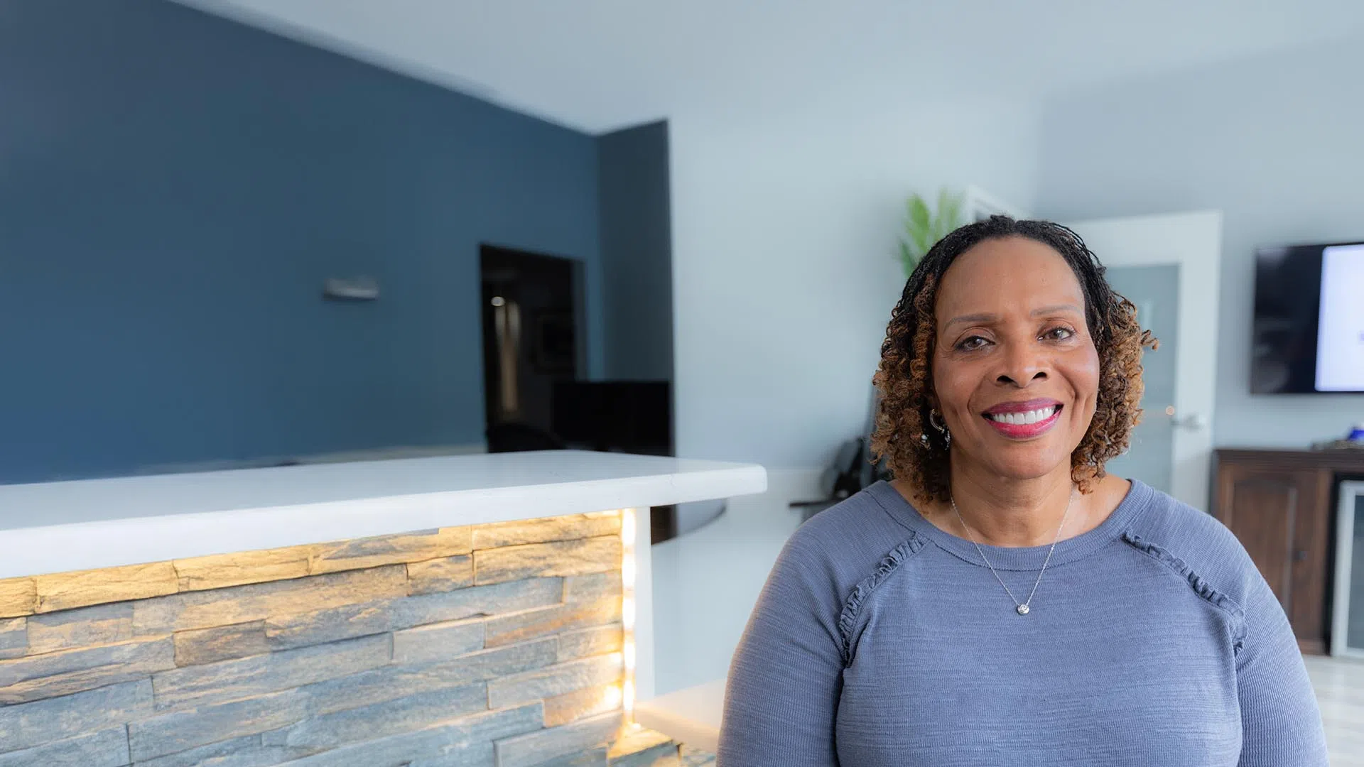 Smiling middle-aged woman with curly hair sitting in a modern living room with blue walls and stone counter.