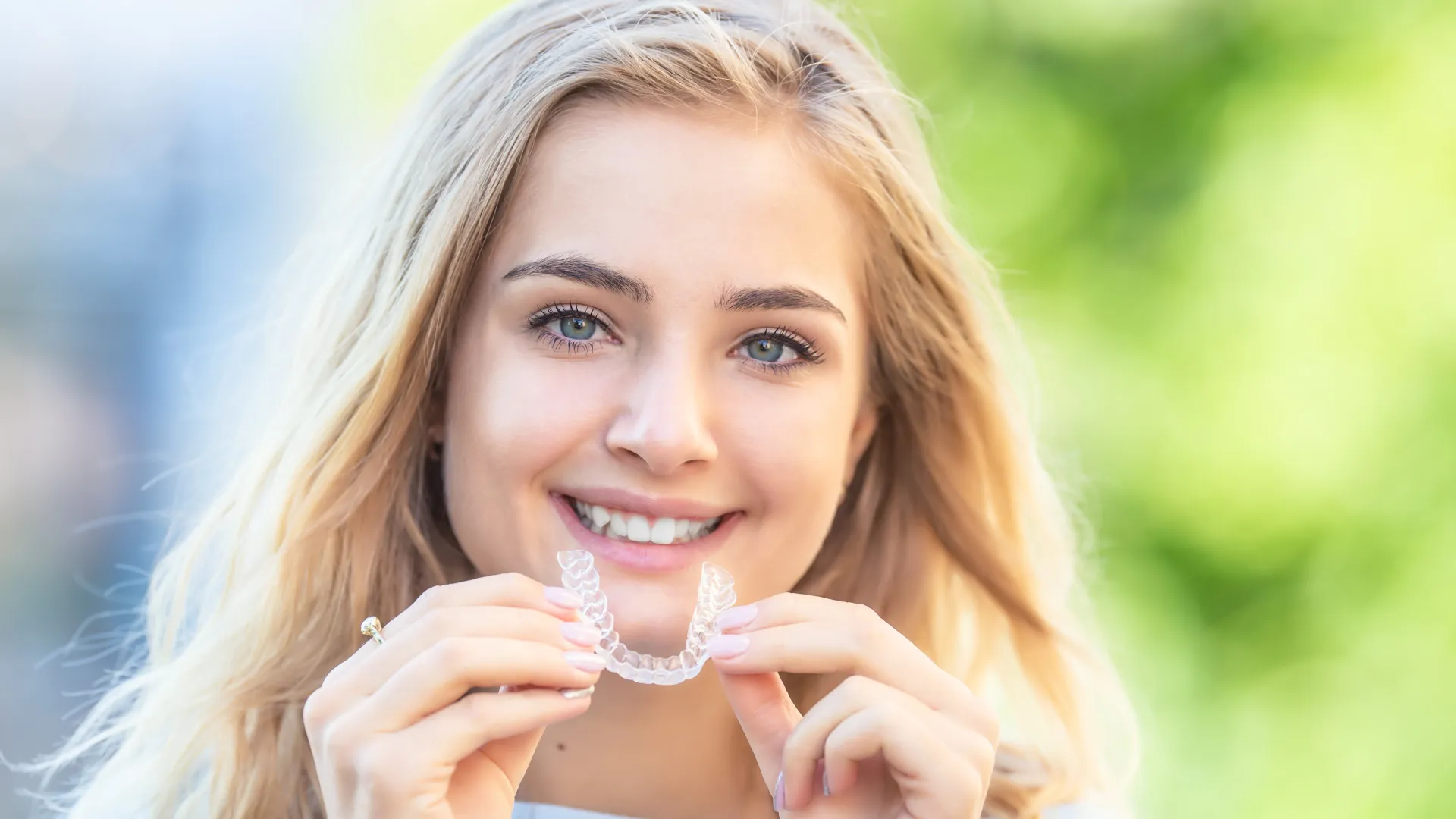 Smiling young woman holding a clear dental aligner outdoors with a blurred green background