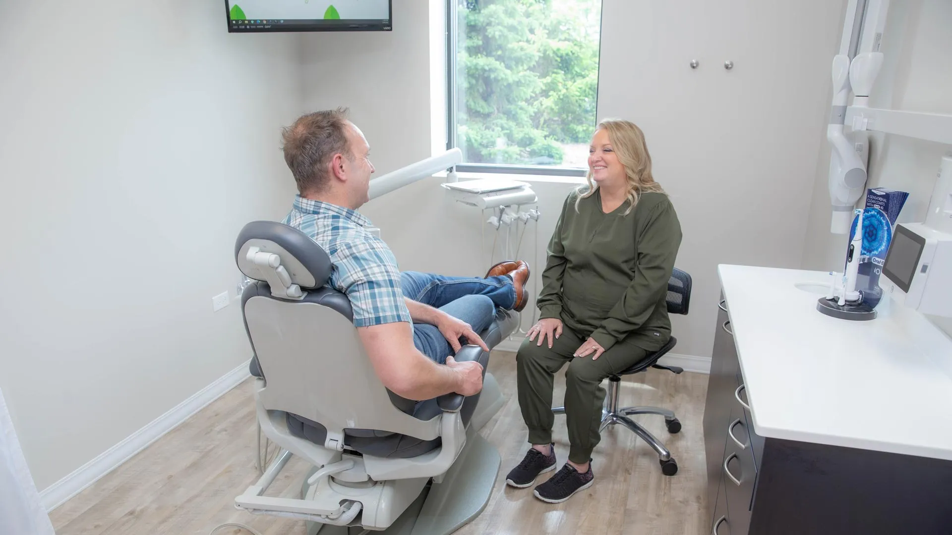 Dentist and patient smiling and talking in a bright, modern dental office with large window and equipment.