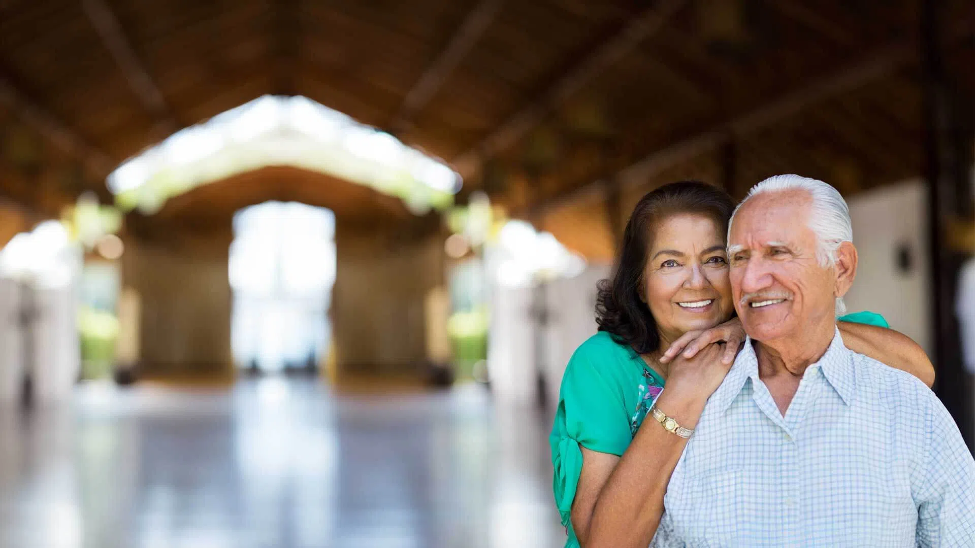 Smiling elderly couple embracing in a bright, spacious indoor hallway with natural light in the background